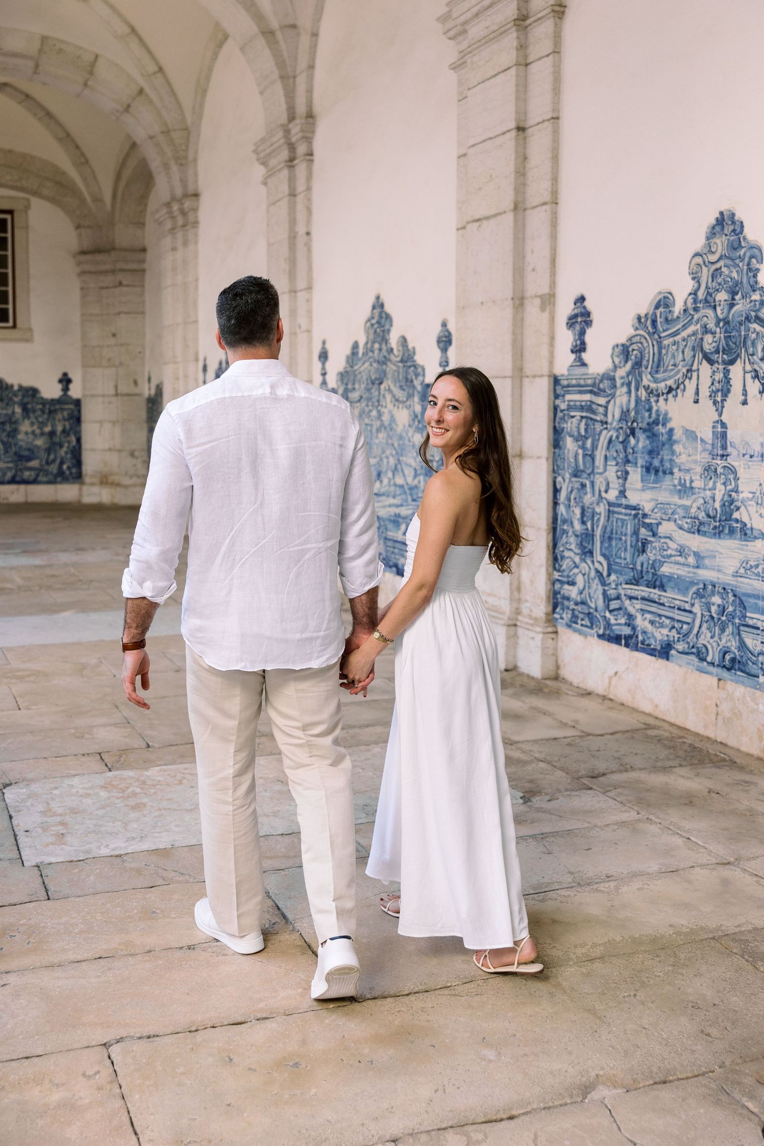 Couple holding hands walking past ornate blue azulejo tile wall in historic Portuguese building.