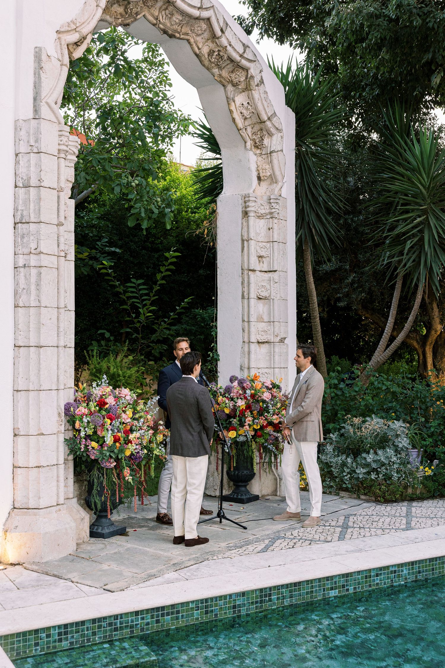 Three men gather under an ornate stone arch adorned with colorful floral arrangements beside a pool.