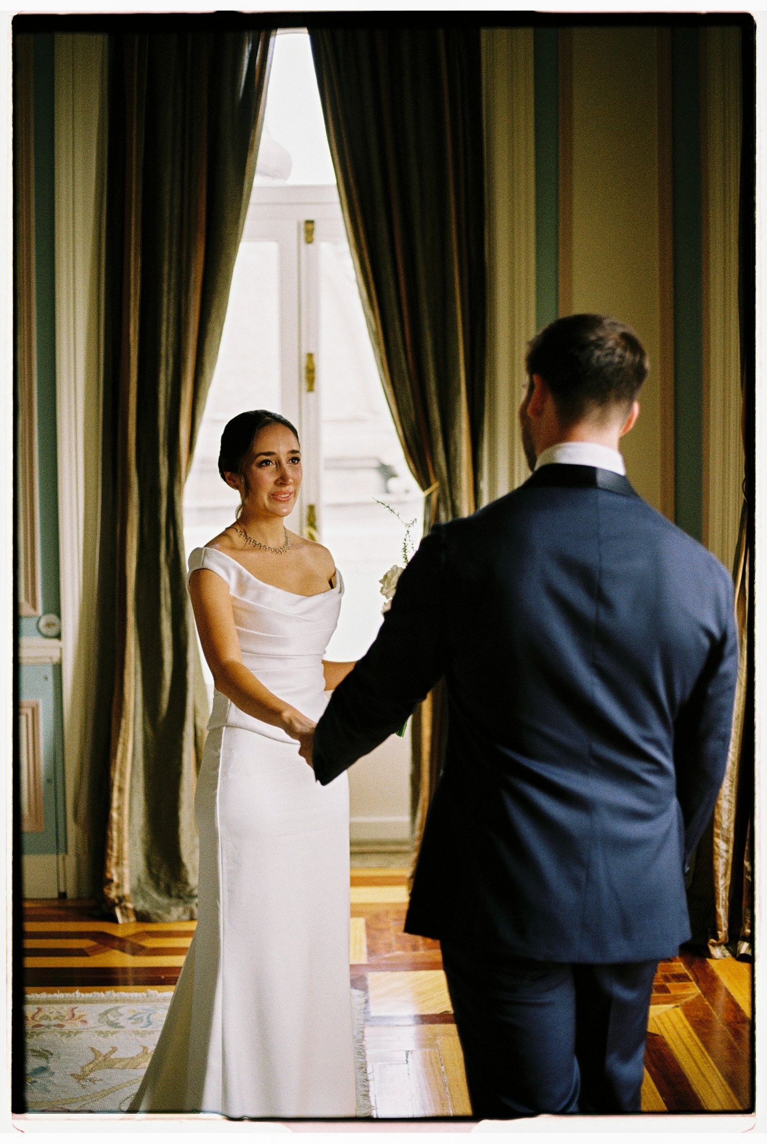 Bride and groom hold hands in elegant room with tall windows and green drapes during wedding first look.