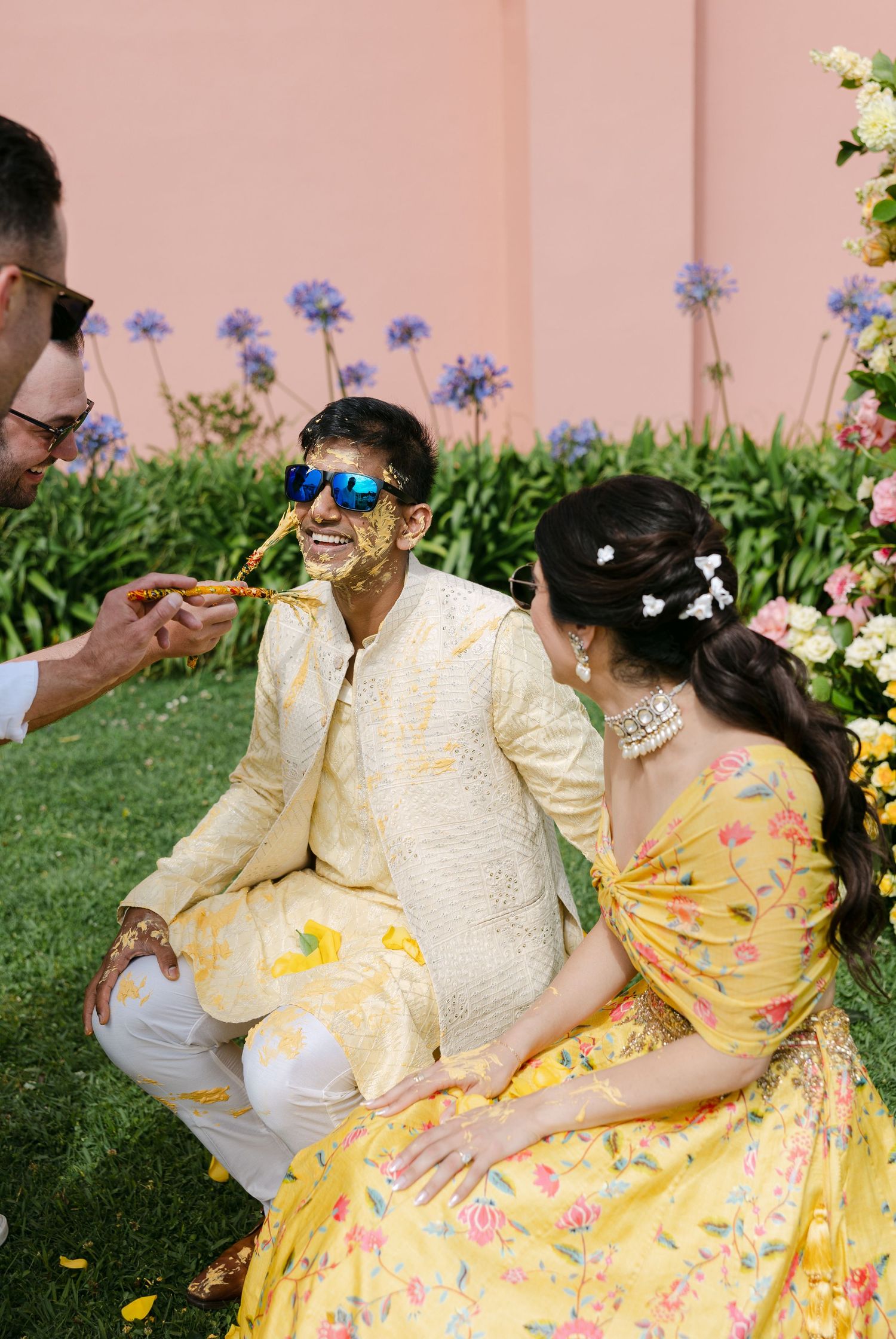 Couple in traditional wedding attire sharing a romantic moment among purple flowers in a garden setting.