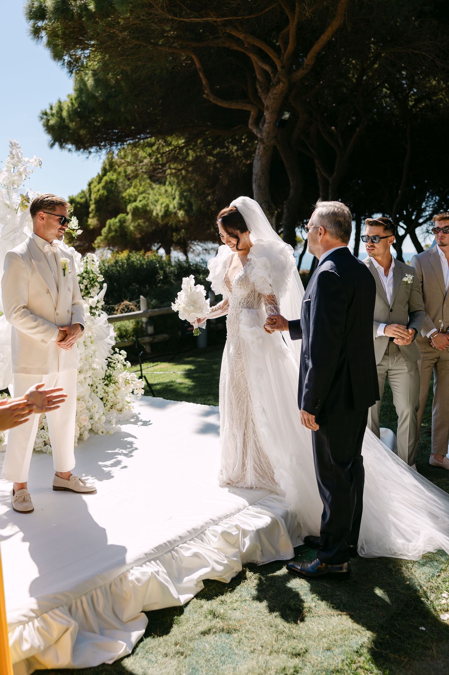 Outdoor wedding ceremony with couple and guests on white platform surrounded by trees.