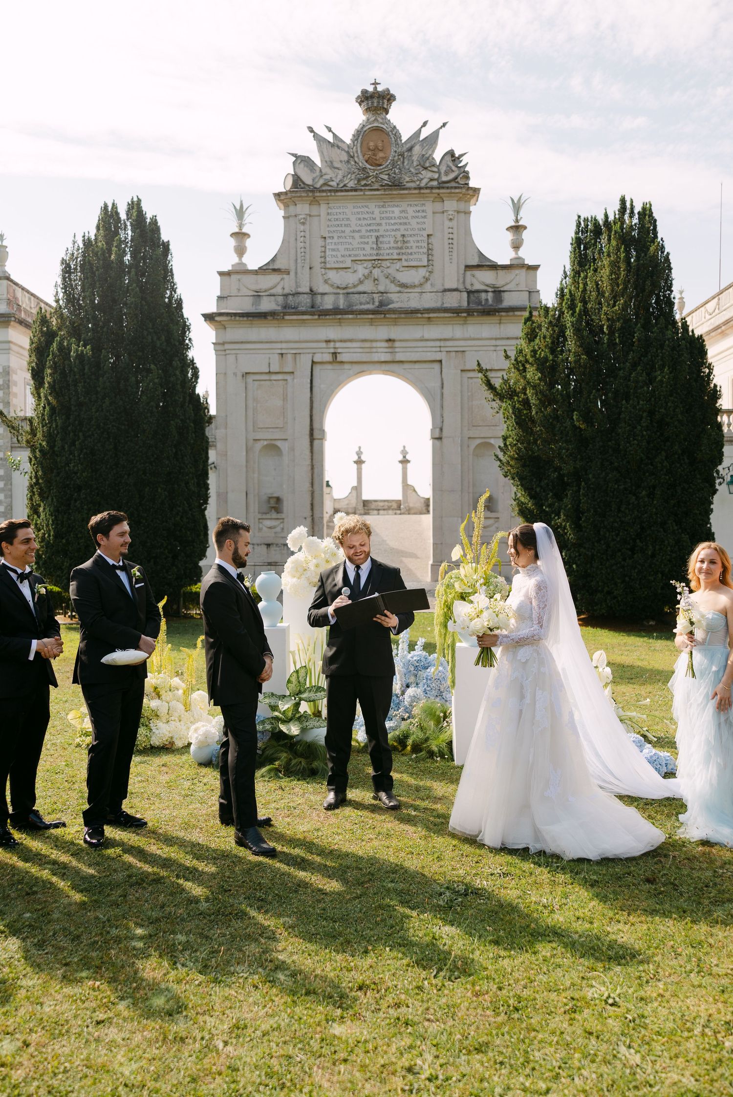 Wedding party stands in front of ornate white stone archway during outdoor ceremony.