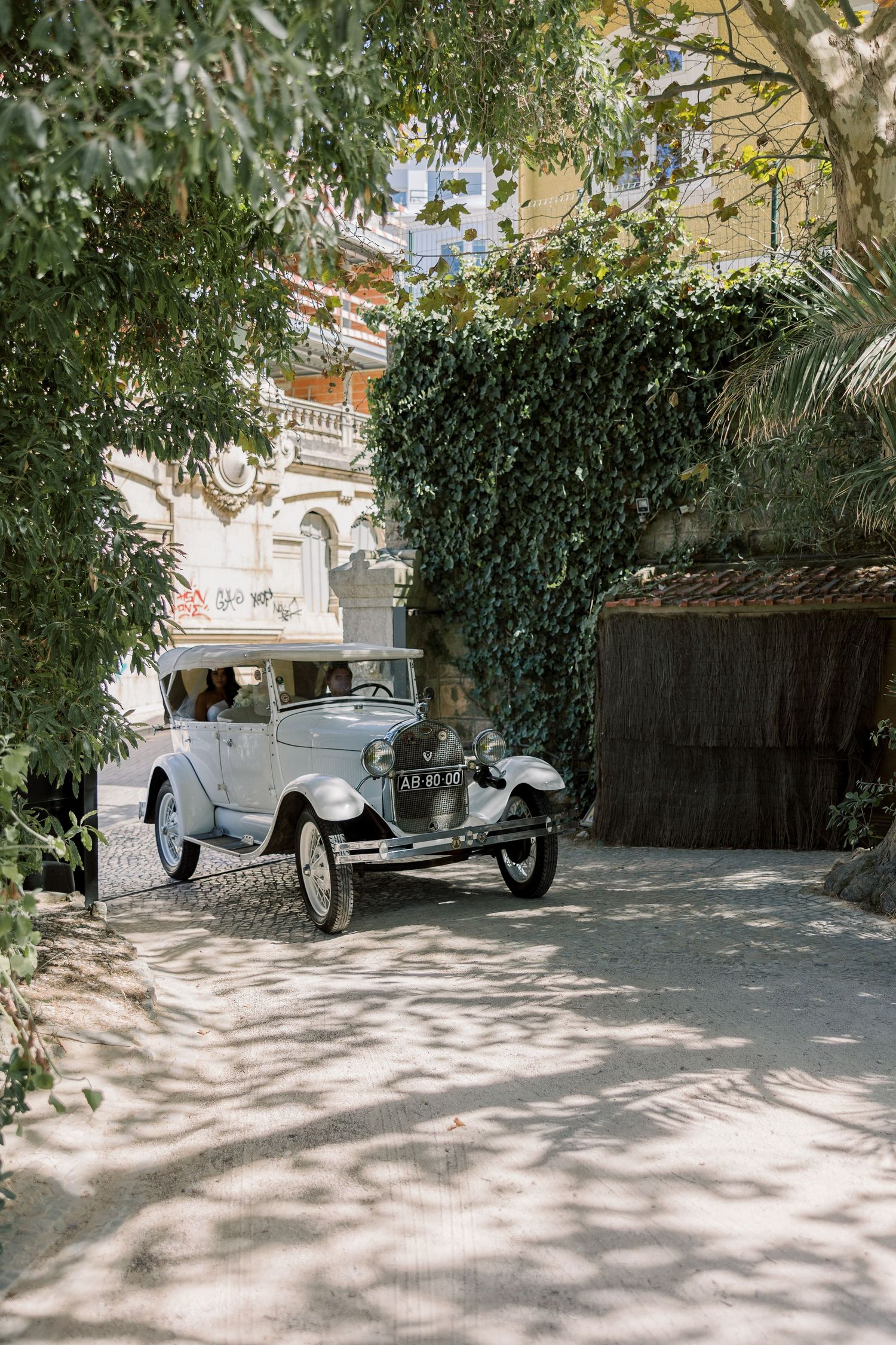 Vintage white car parked in a shaded driveway with trees and buildings in the background.