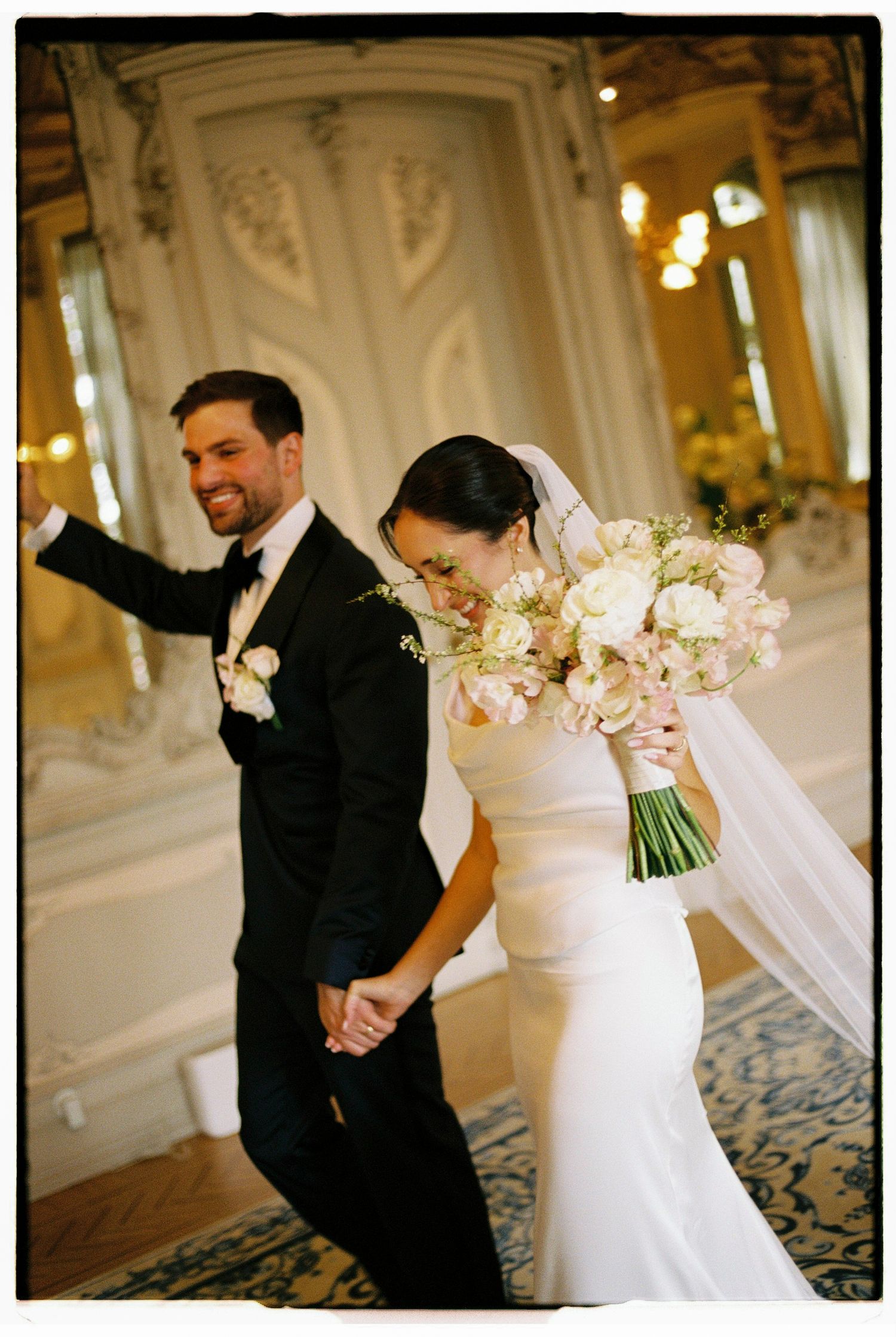 Smiling groom in tuxedo holds hands with bride in white gown and veil holding floral bouquet in elegant ballroom.