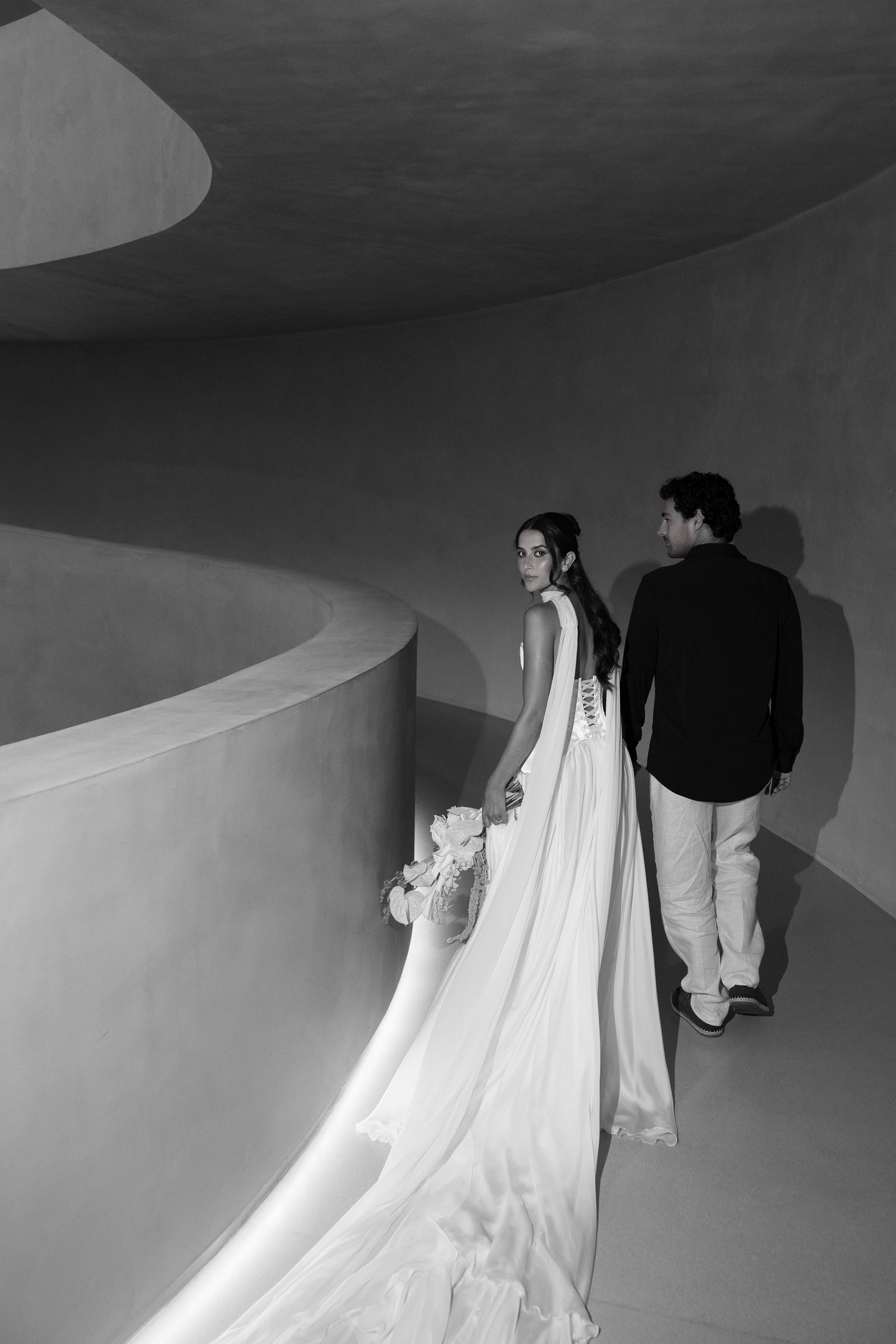 Black and white photo of bride in long veil and groom walking through curved architectural corridor.