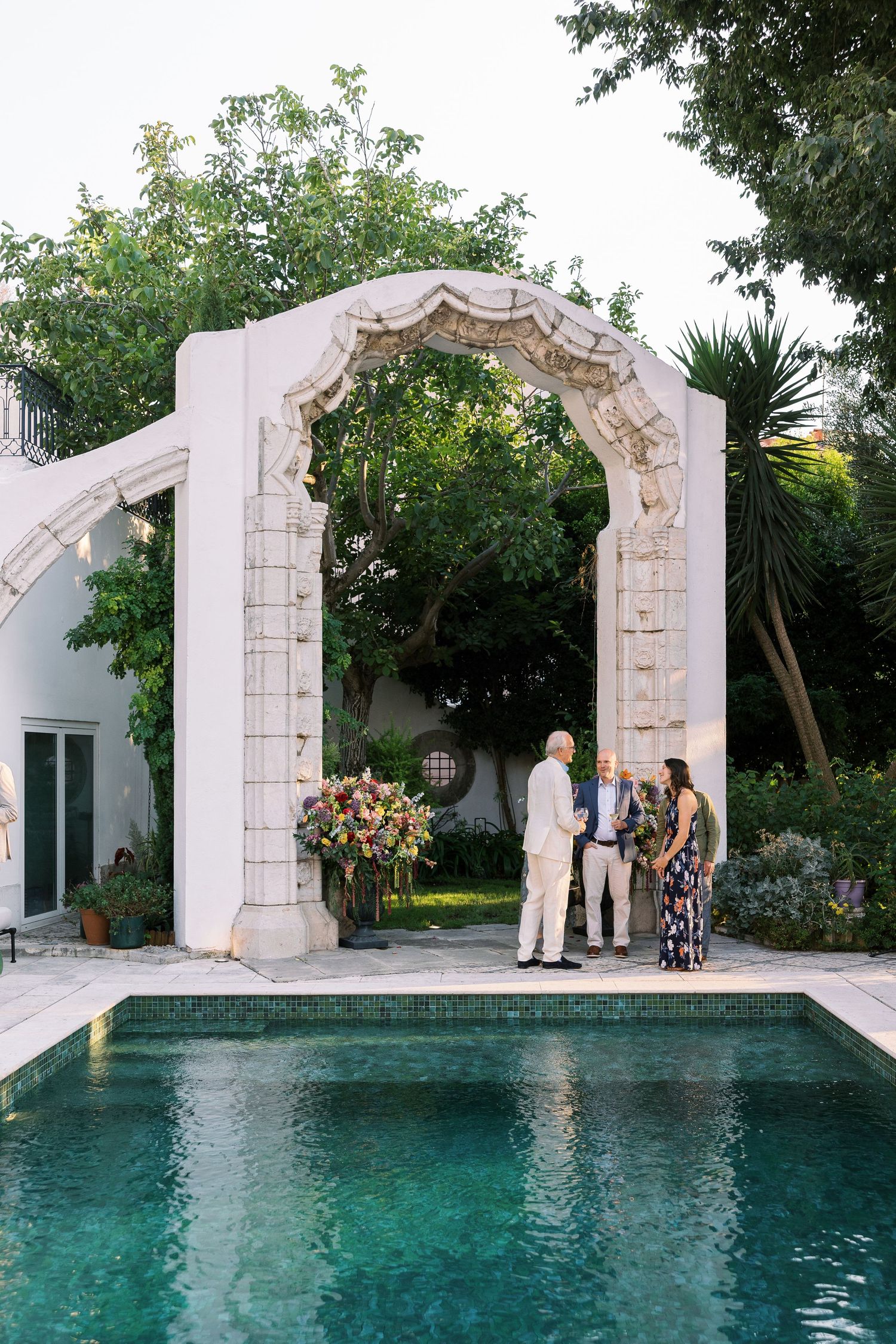Intimate outdoor wedding ceremony beside a pool under an ornate white stone arch surrounded by lush greenery.
