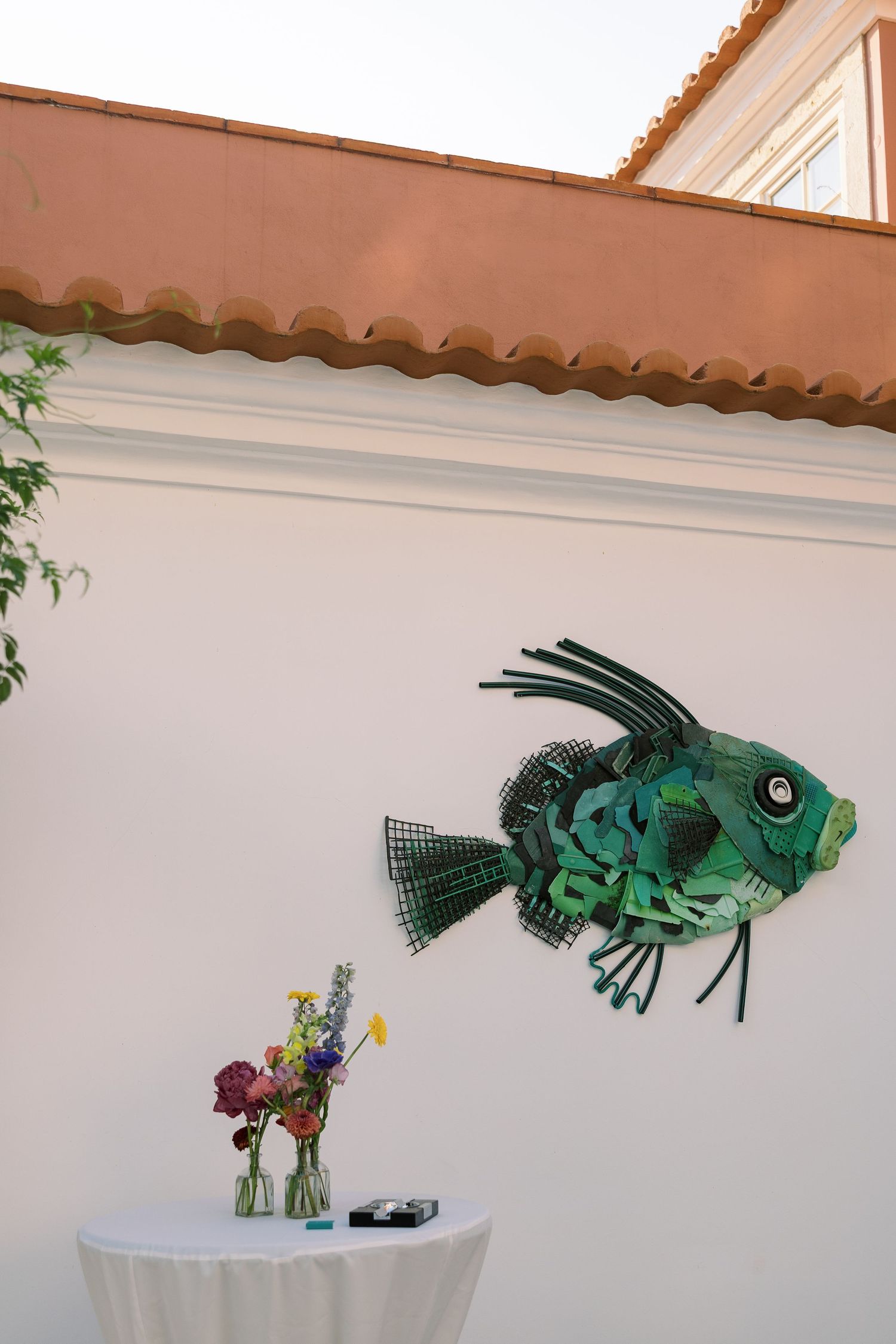 Green fish sculpture made from recycled materials mounted on white wall above a small table with colorful flowers.