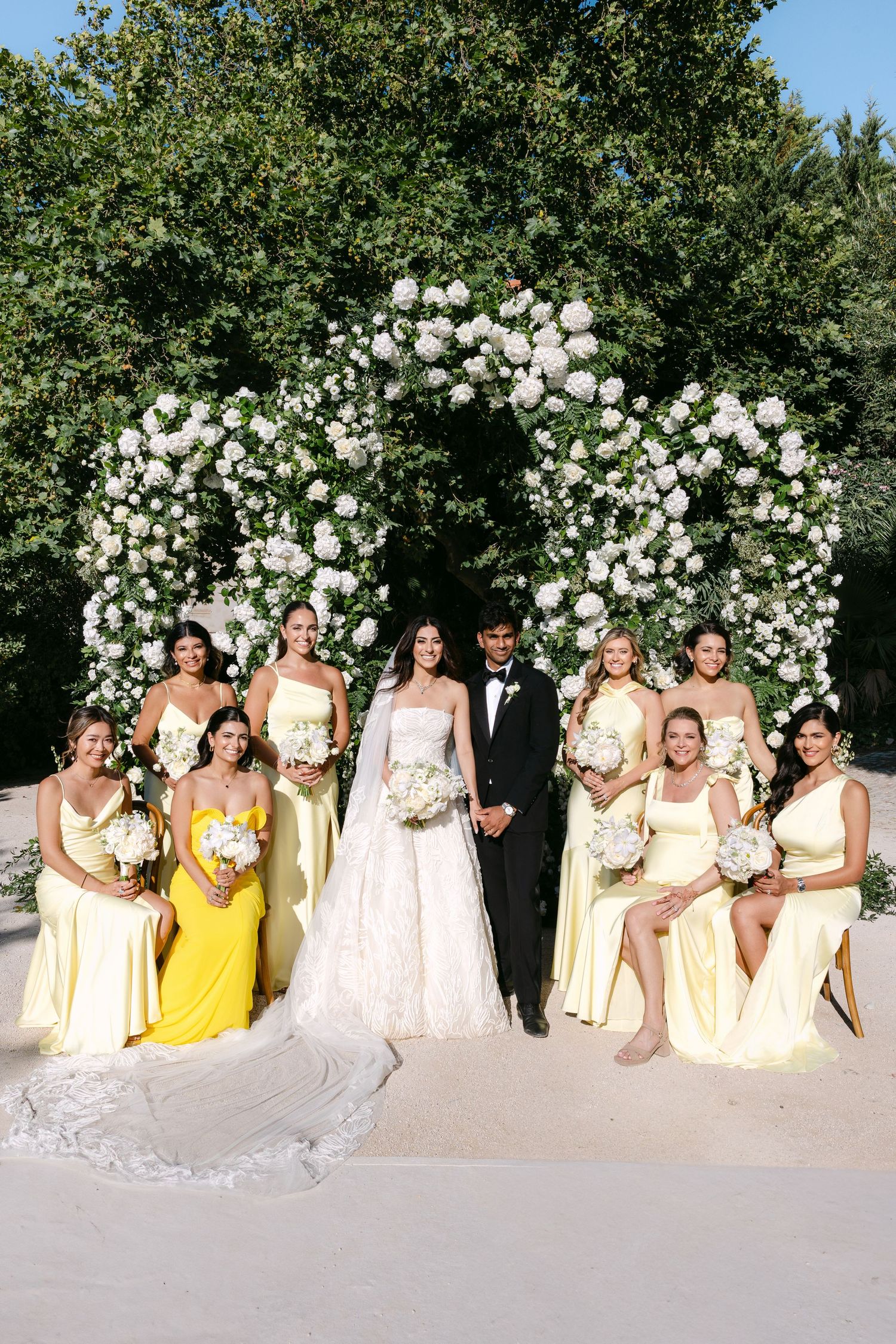 Wedding party in yellow and white dresses posing in front of a floral arch.