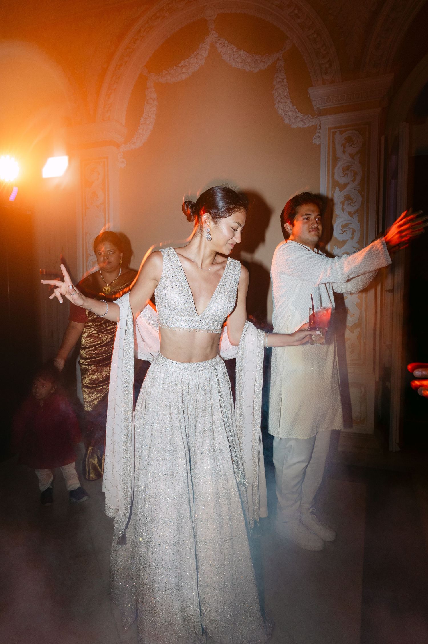 Guests dancing in an ornate ballroom with dramatic lighting and white formal attire.