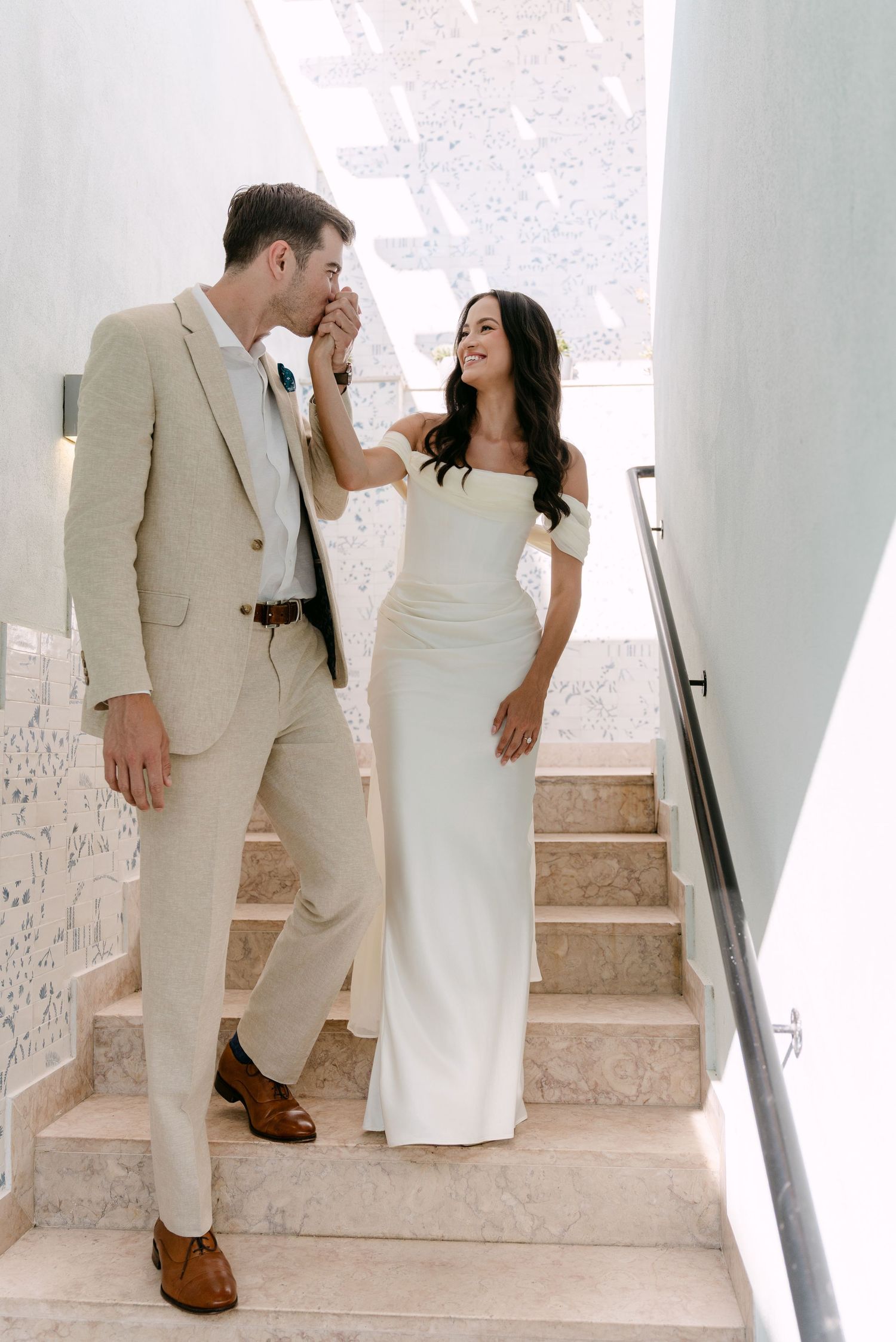 A couple in wedding attire shares a playful moment on white stone stairs.