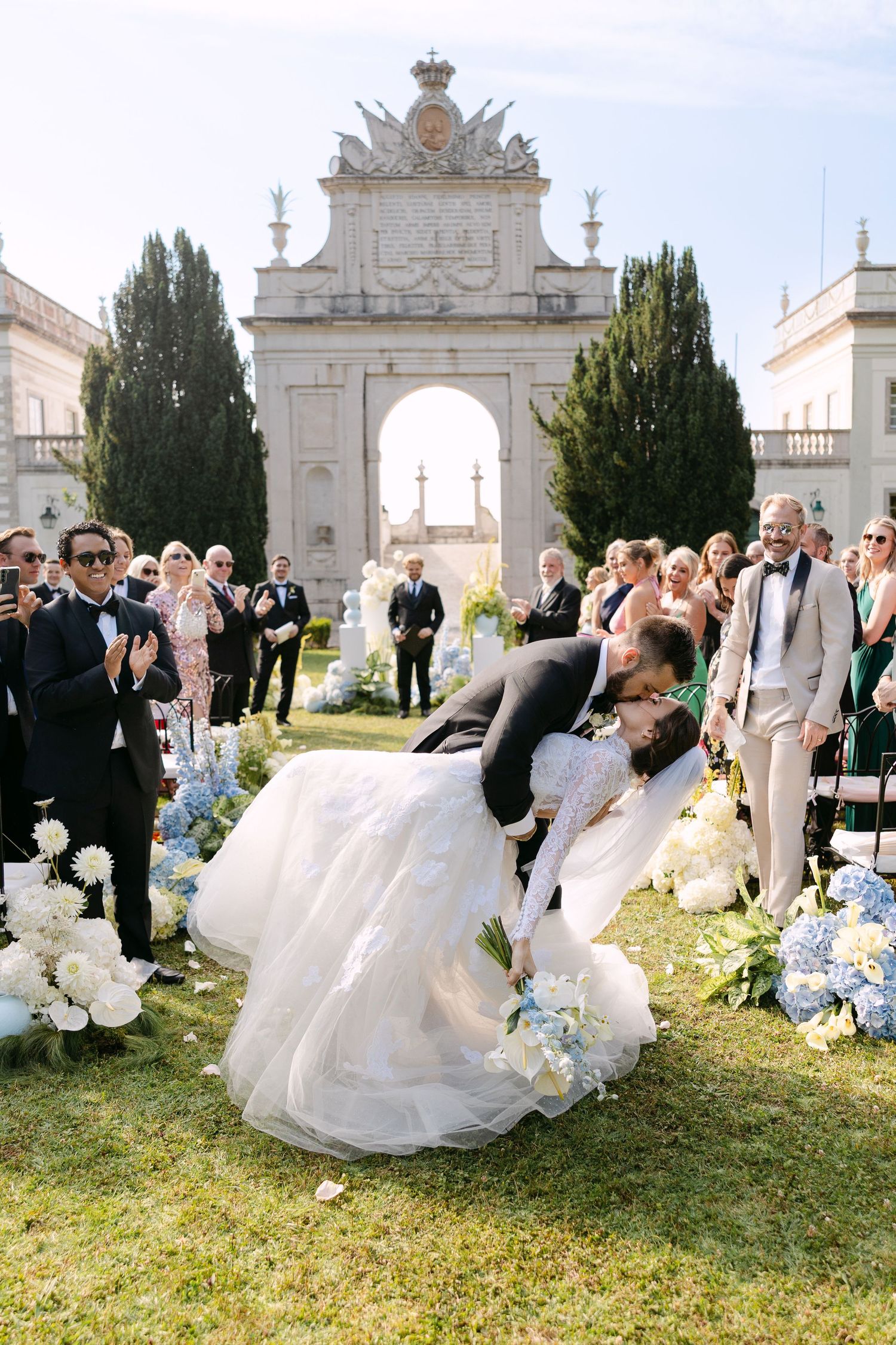 Wedding celebration in front of ornate white stone archway with blue and white floral decorations.