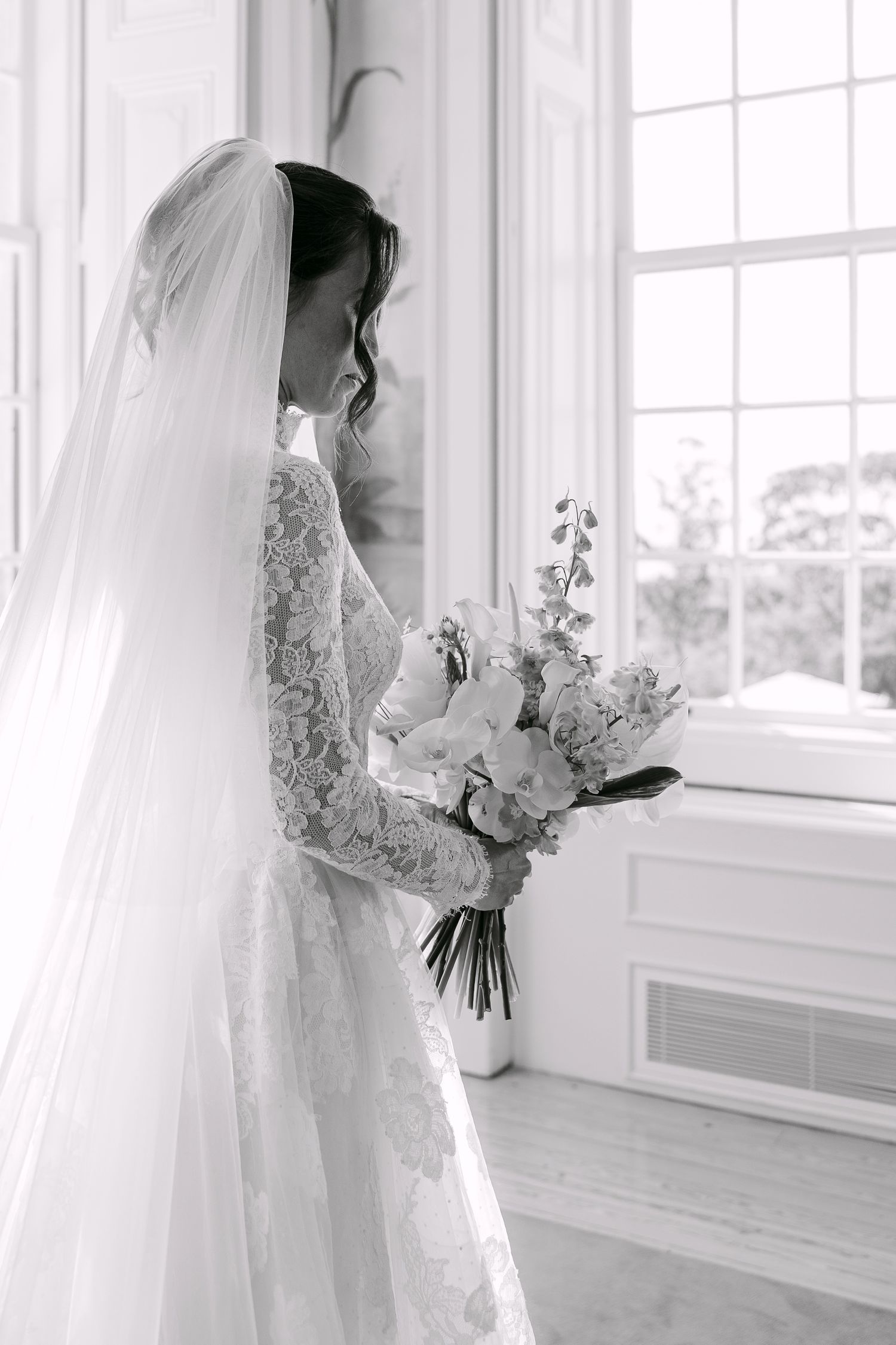 A bride in a lace wedding gown holds a bouquet while standing by a window.