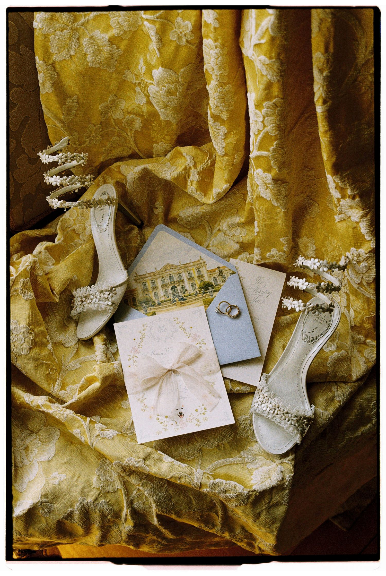 Elegant wedding flat lay with jeweled bridal heels, gold fabric, rings, and floral stationery on golden damask backdrop.