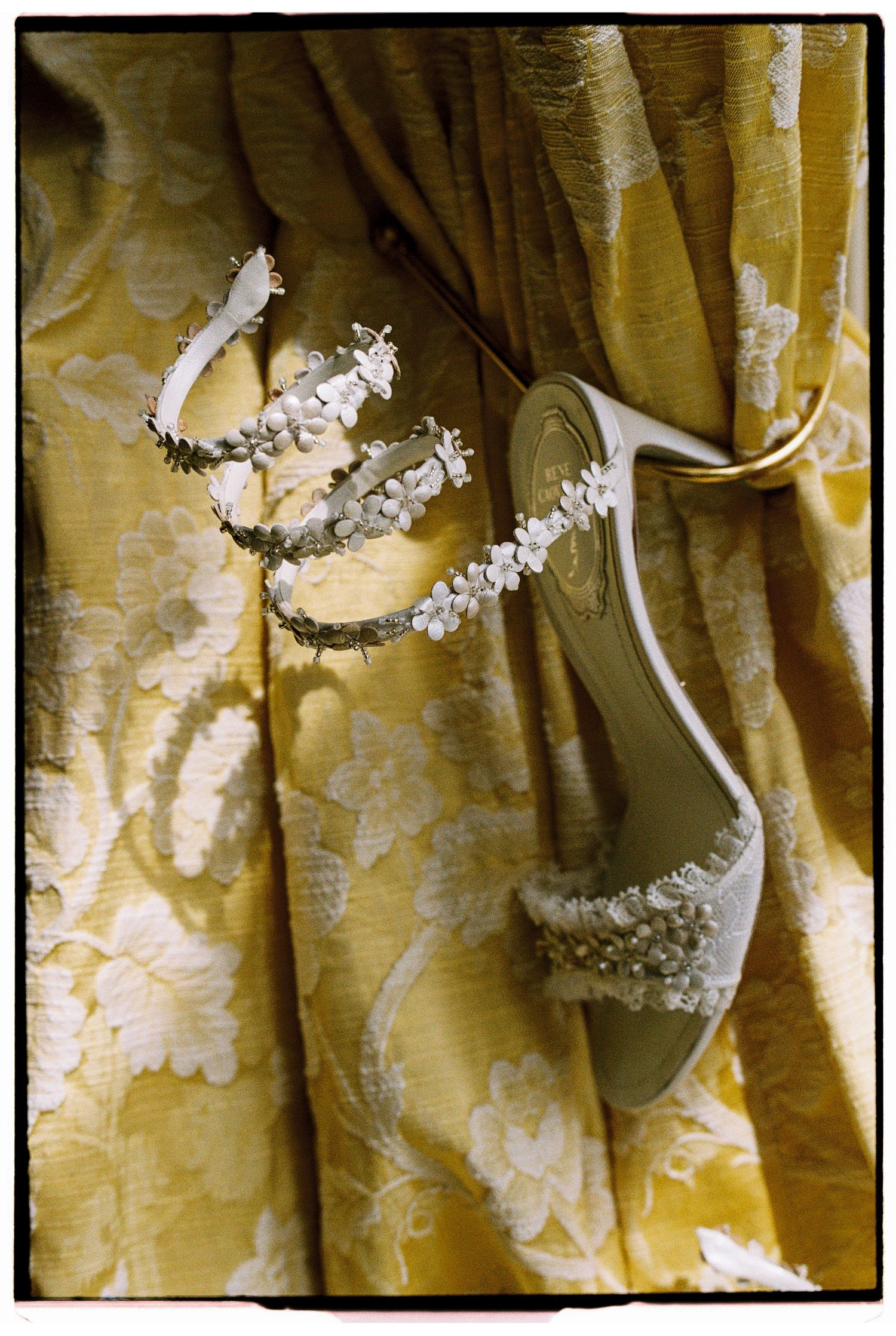 Elegant white floral embellished bridal heels displayed against golden damask fabric curtain.