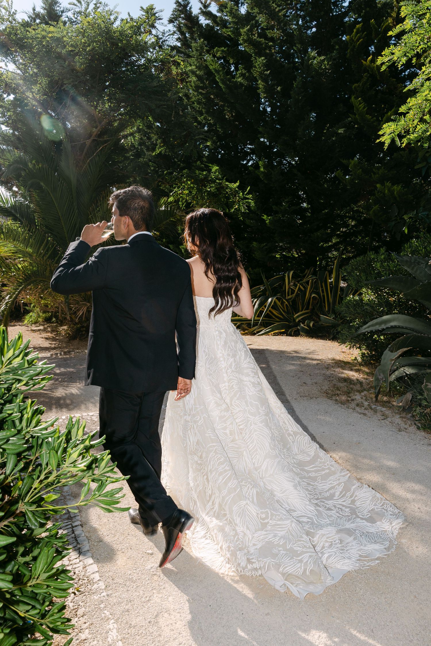 Long flowing wedding dress train trailing behind couple walking through garden path.
