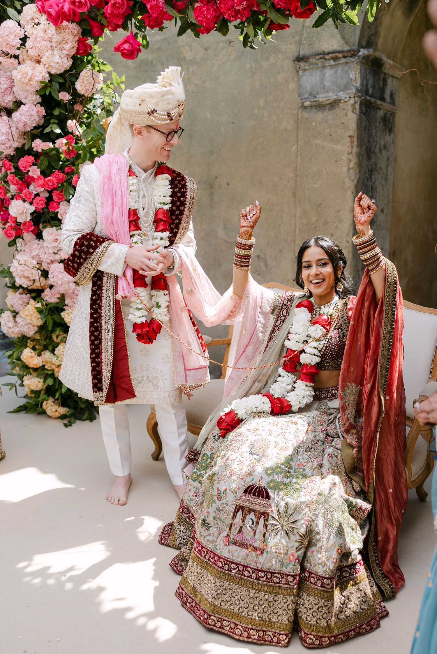 Couple performs traditional wedding rituals under a floral arch with pink and red roses.