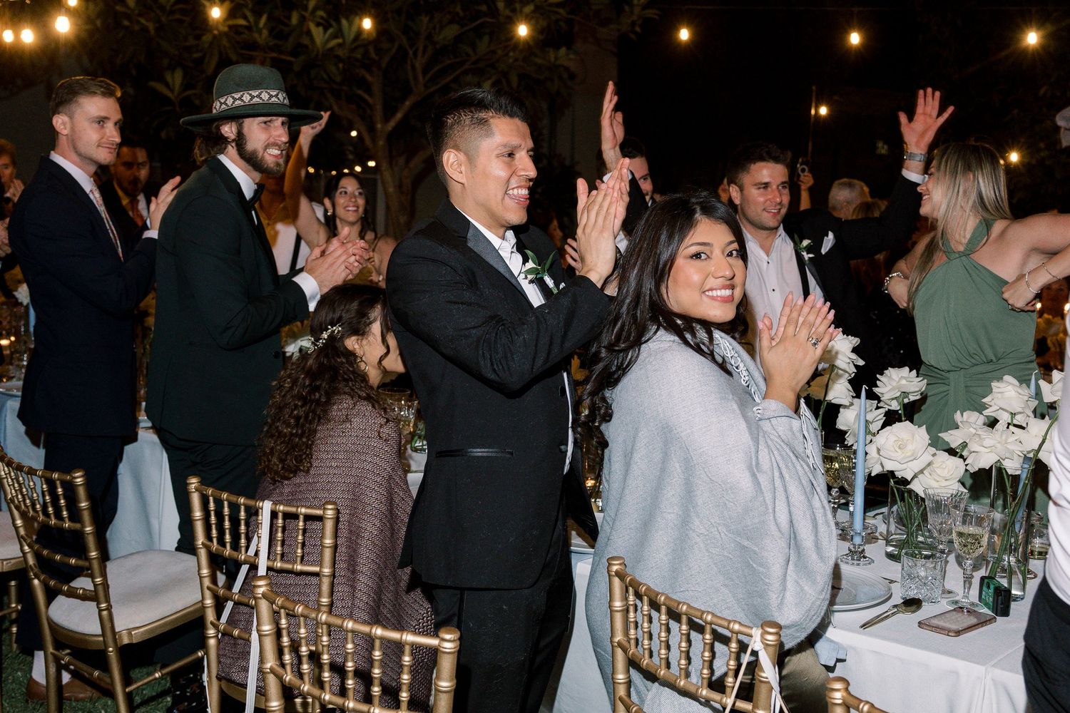 Wedding celebration at night with guests dancing and cheering under string lights.