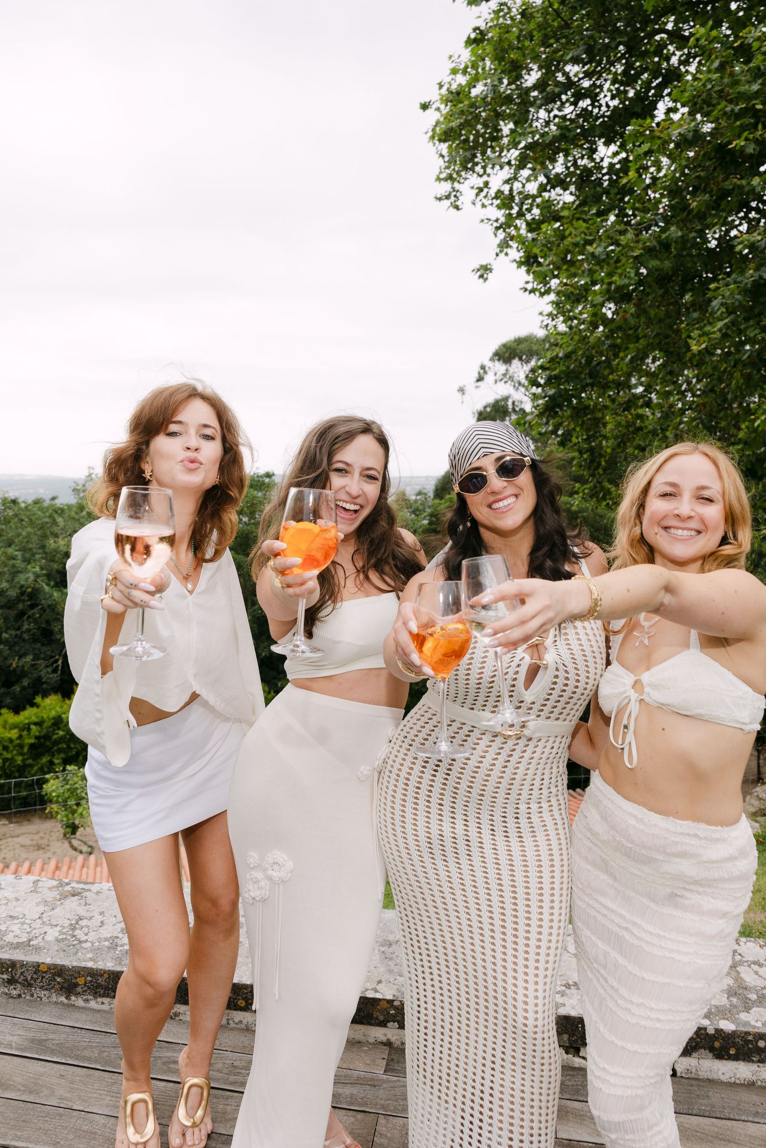 Four friends in white outfits toast with cocktails on an outdoor terrace.