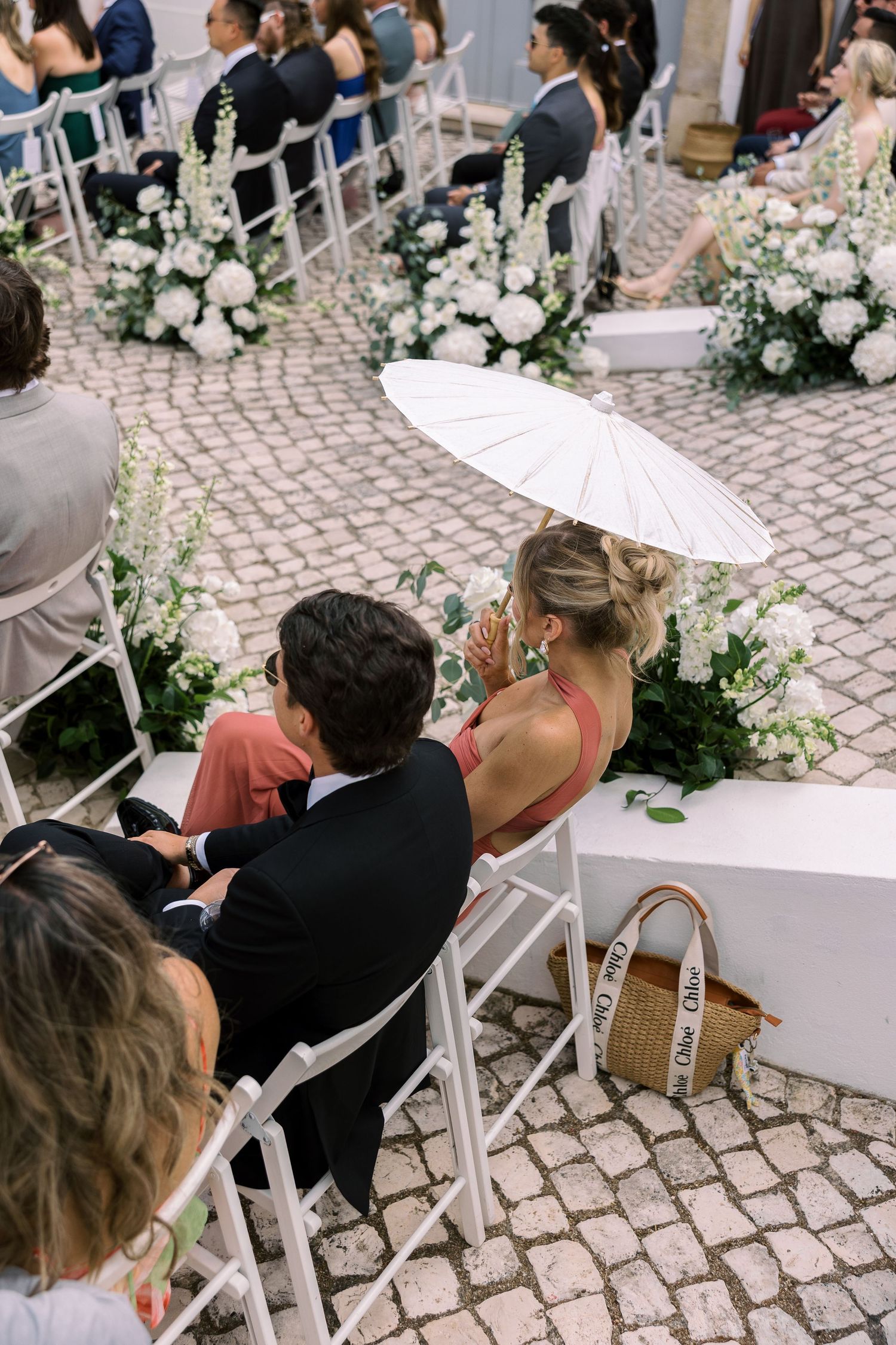 Guests seated at an outdoor wedding ceremony with white floral arrangements, a woman holding a white parasol umbrella.