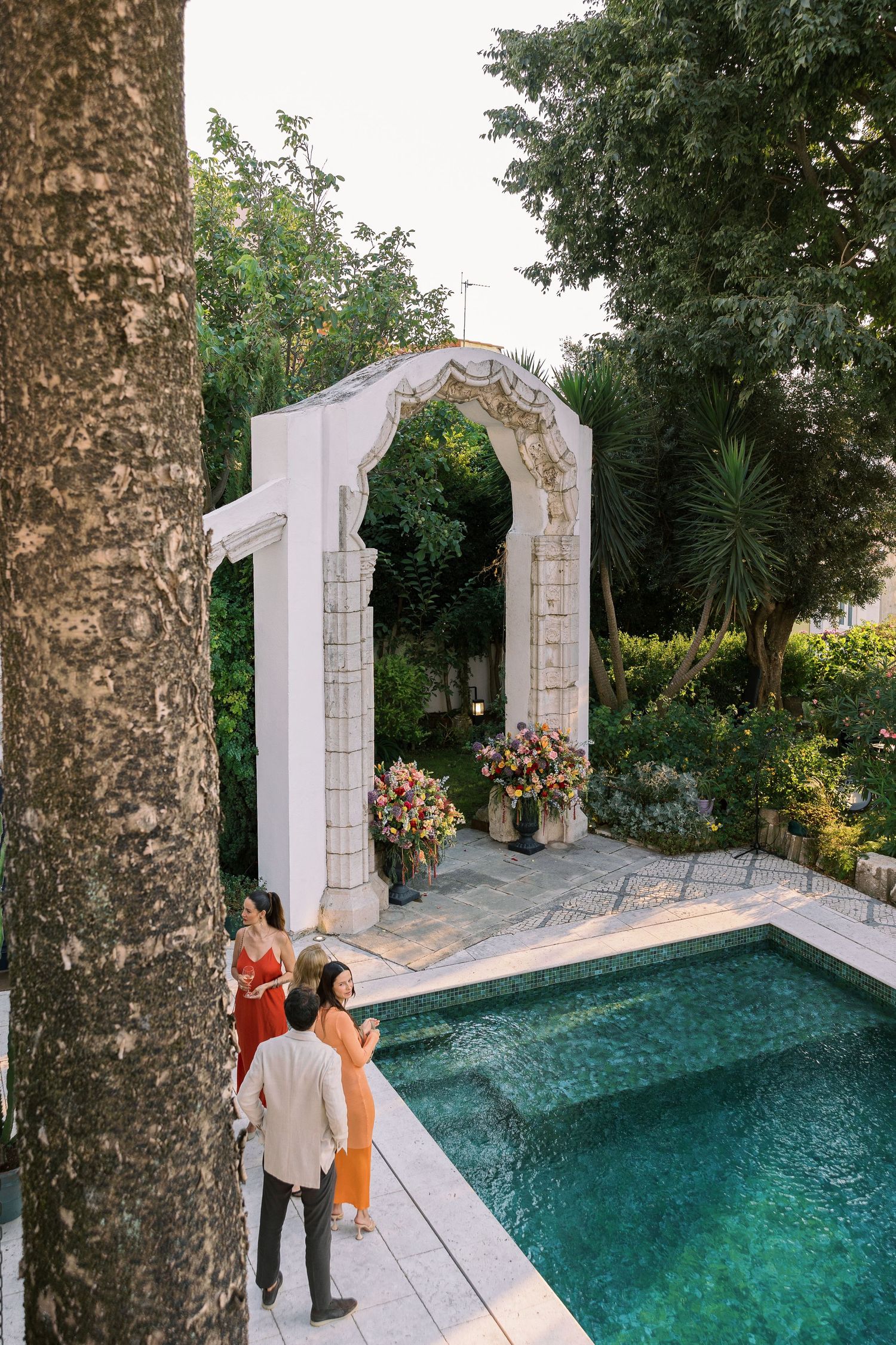 Elegant outdoor wedding arch by a pool surrounded by lush gardens with guests in colorful attire.