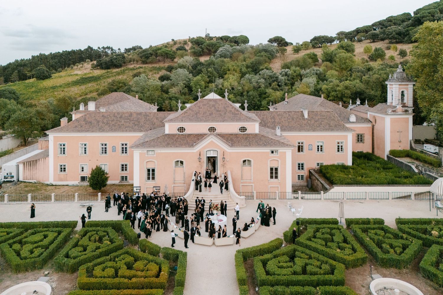 Aerial view of a pink Mediterranean-style estate with formal gardens and gathered wedding guests.