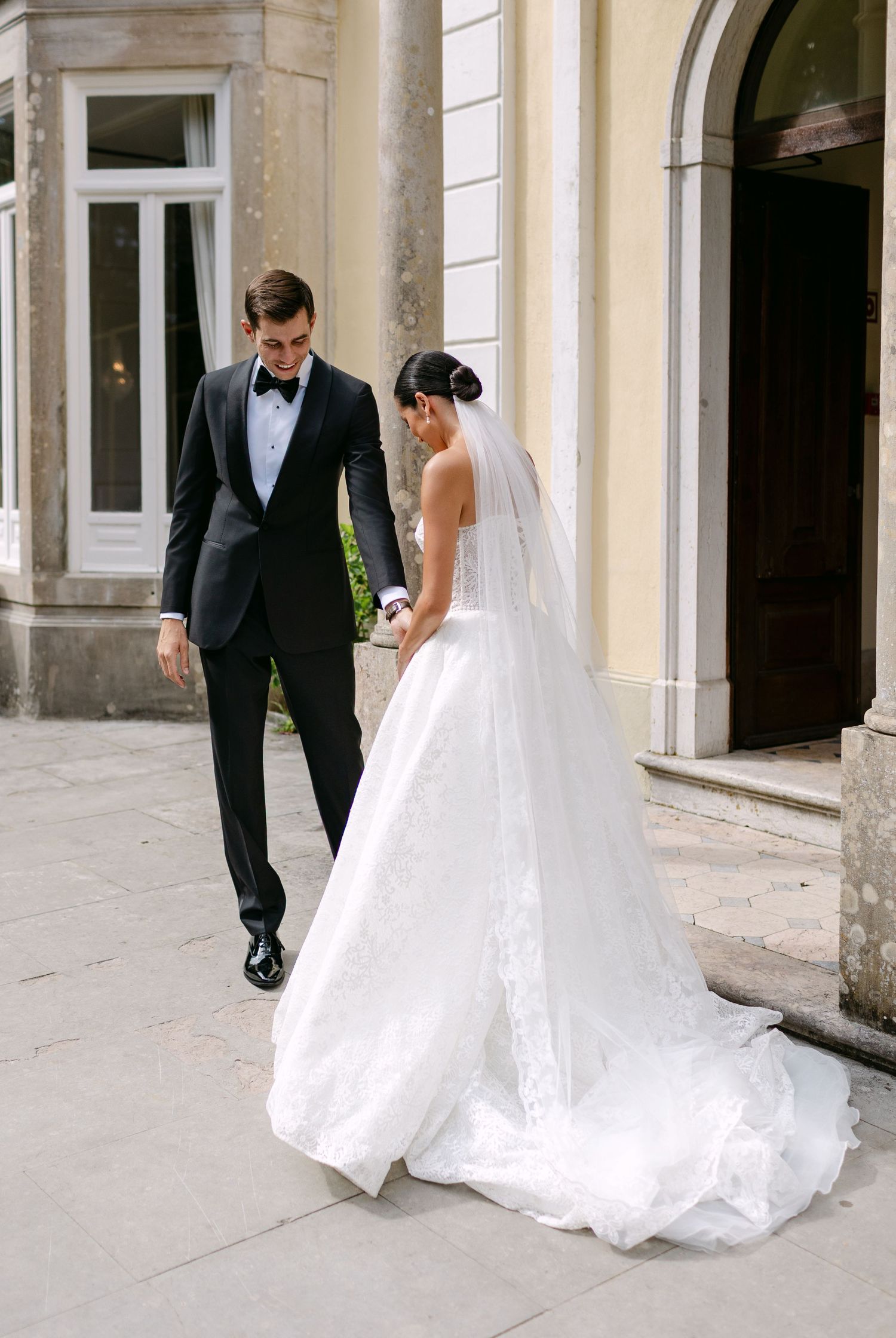 Couple in wedding attire walking on stone patio outside classical building entrance.