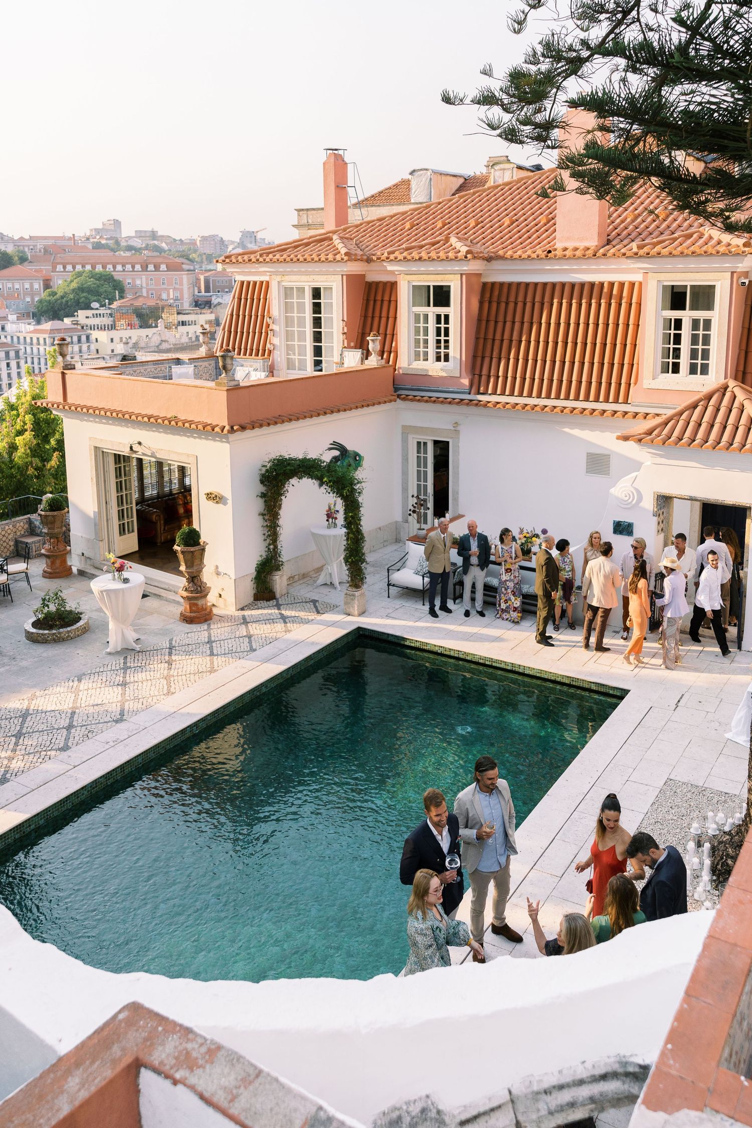 Elegant outdoor wedding reception at a Mediterranean villa with pool, terrace, and terracotta roof tiles overlooking a city.