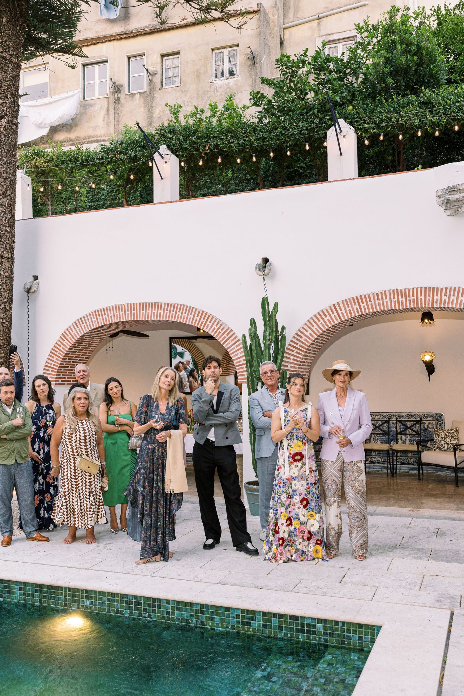 Elegantly dressed wedding guests gather poolside at a luxury villa with string lights and arched walls.