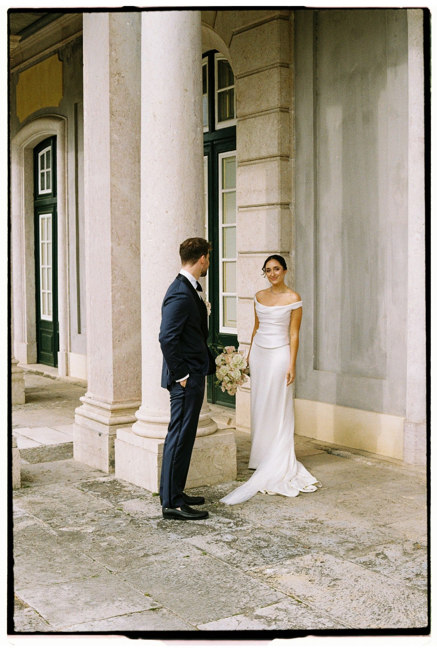 Bride in off-shoulder white gown holds bouquet facing groom in navy suit beside stone columns.