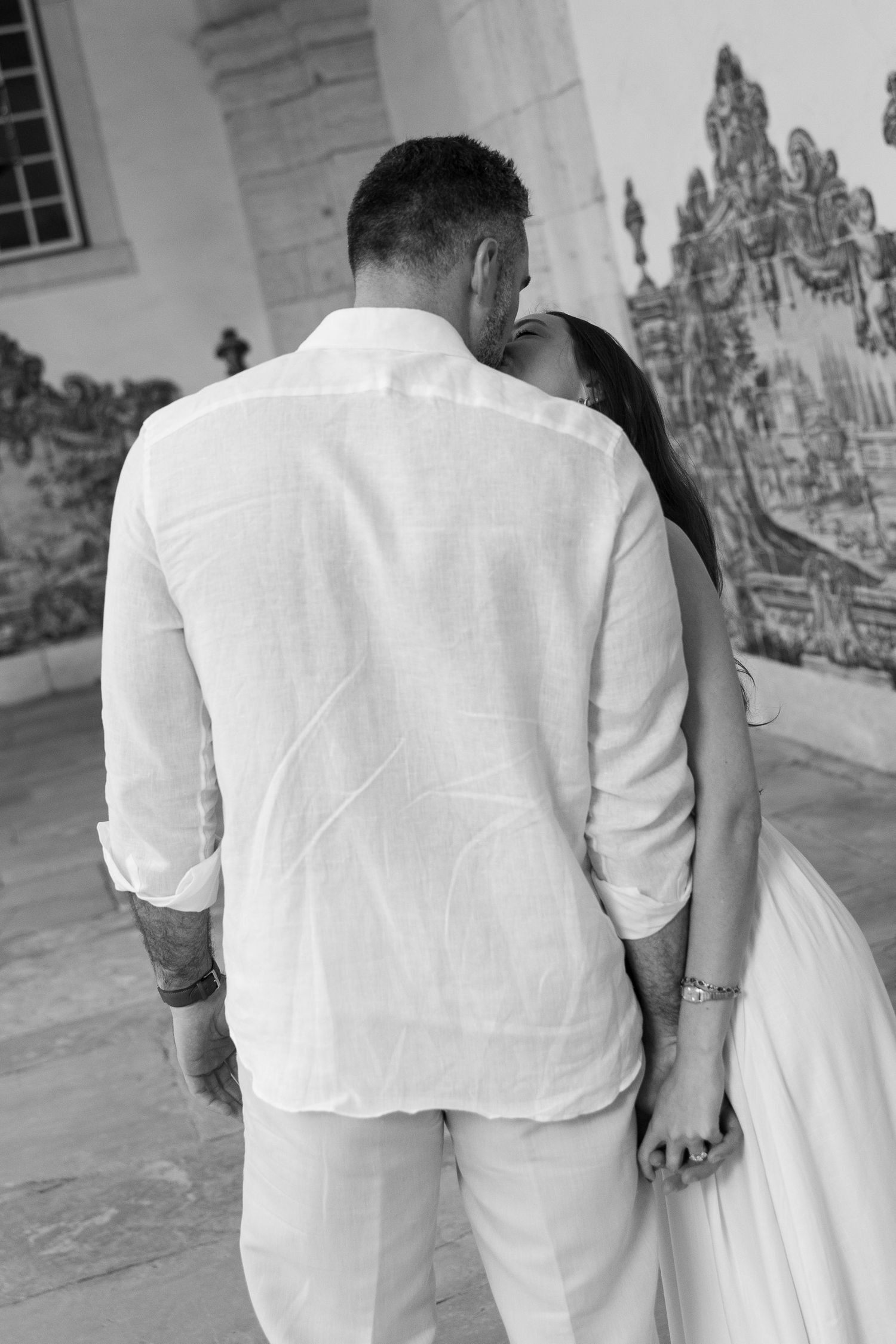 Black and white photo of a couple in white outfits sharing a romantic moment near decorative azulejo tiles.