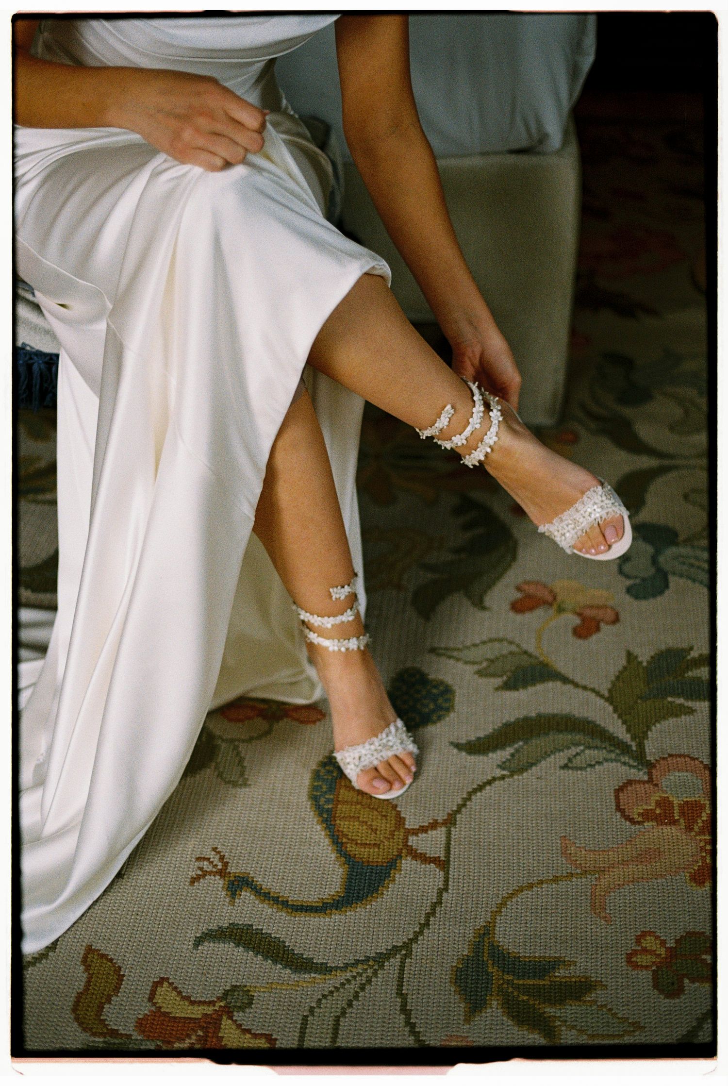 Bride sitting and fastening pearl-embellished strappy wedding sandals over a white satin gown.