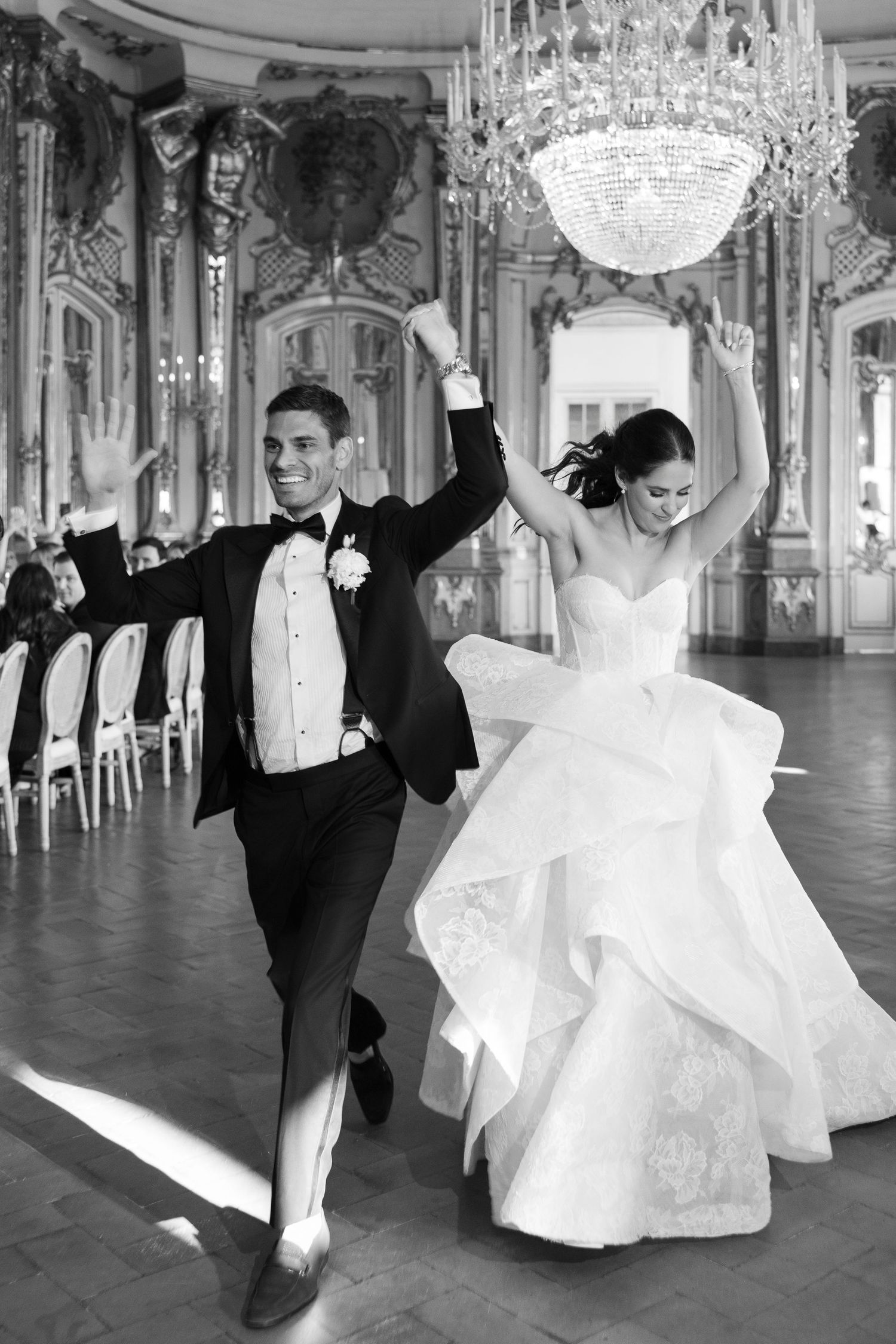 Black and white photo of joyful bride and groom dancing with raised hands in a grand ornate ballroom.