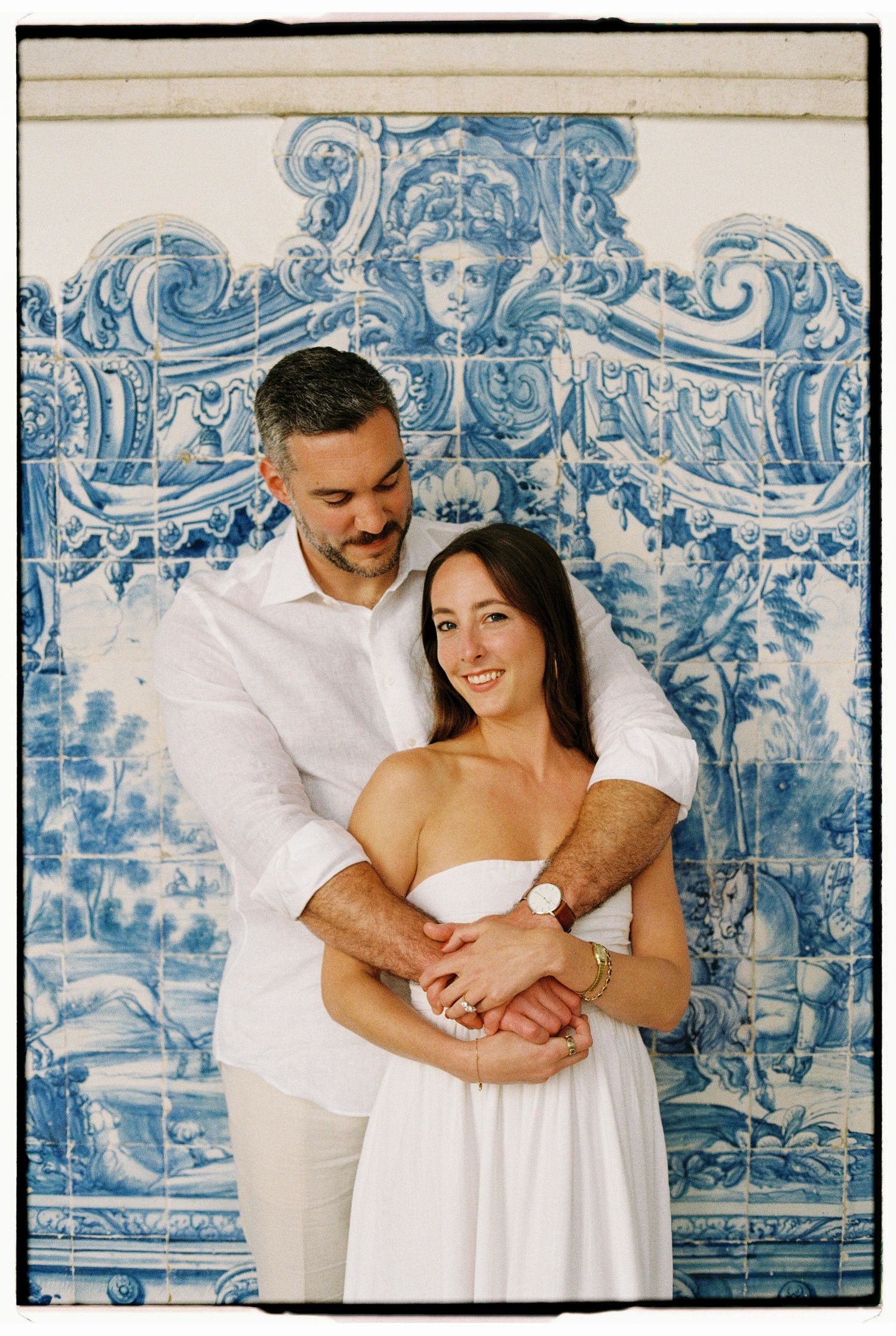 Couple embracing in front of ornate blue and white Portuguese azulejo tile wall, woman in white dress smiling.