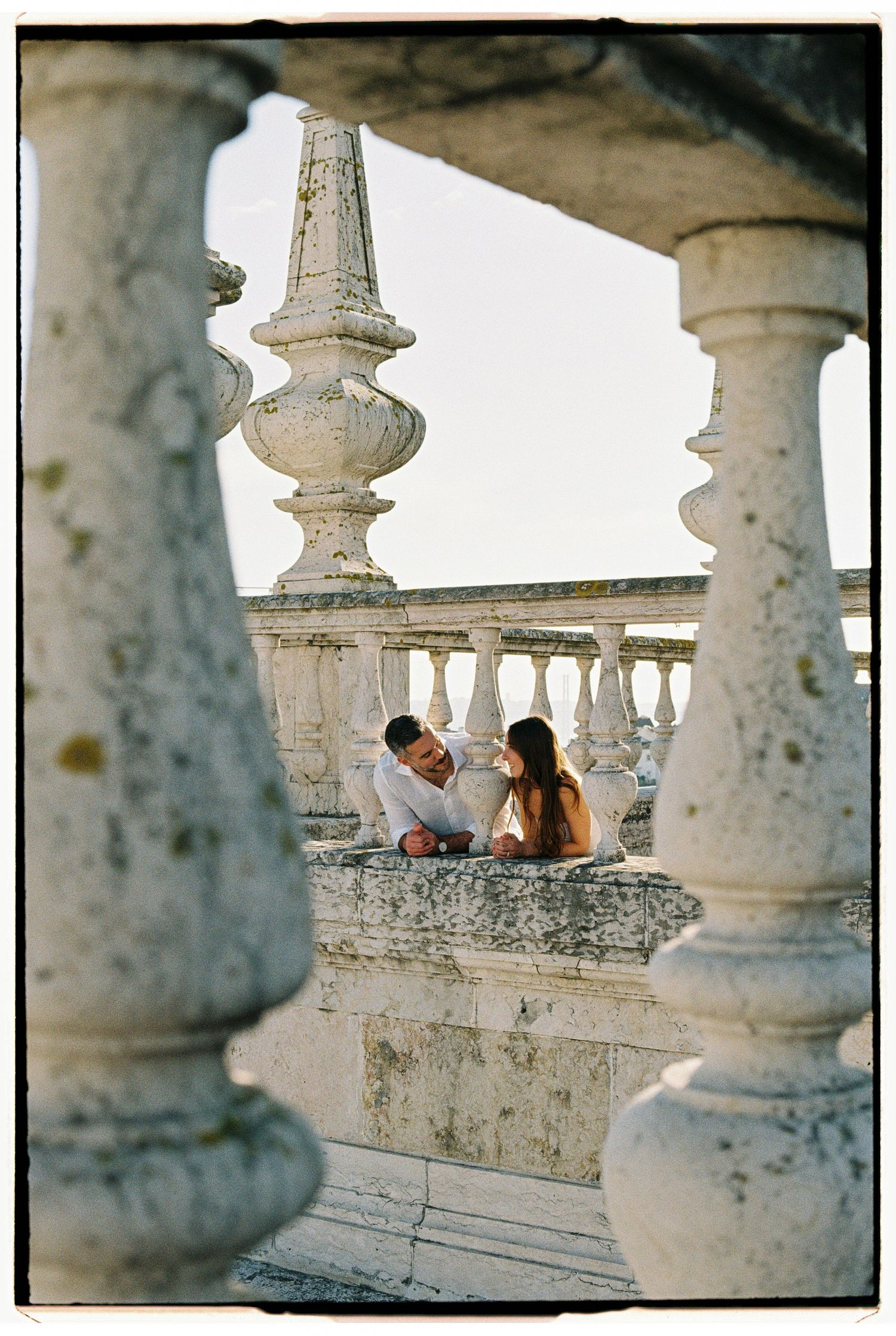 Couple sitting together on ornate white stone balustrade balcony framed by classical columns.