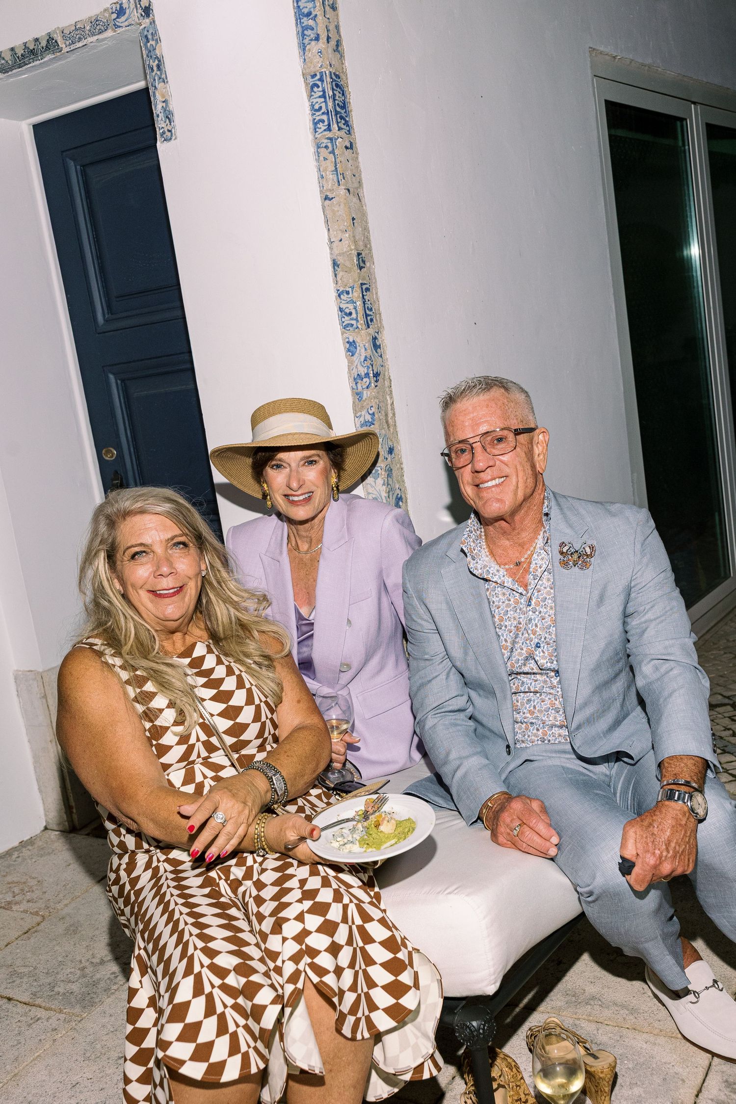Three elegantly dressed guests smile together at an outdoor social event, woman in hat and lavender suit, man in light blue blazer.