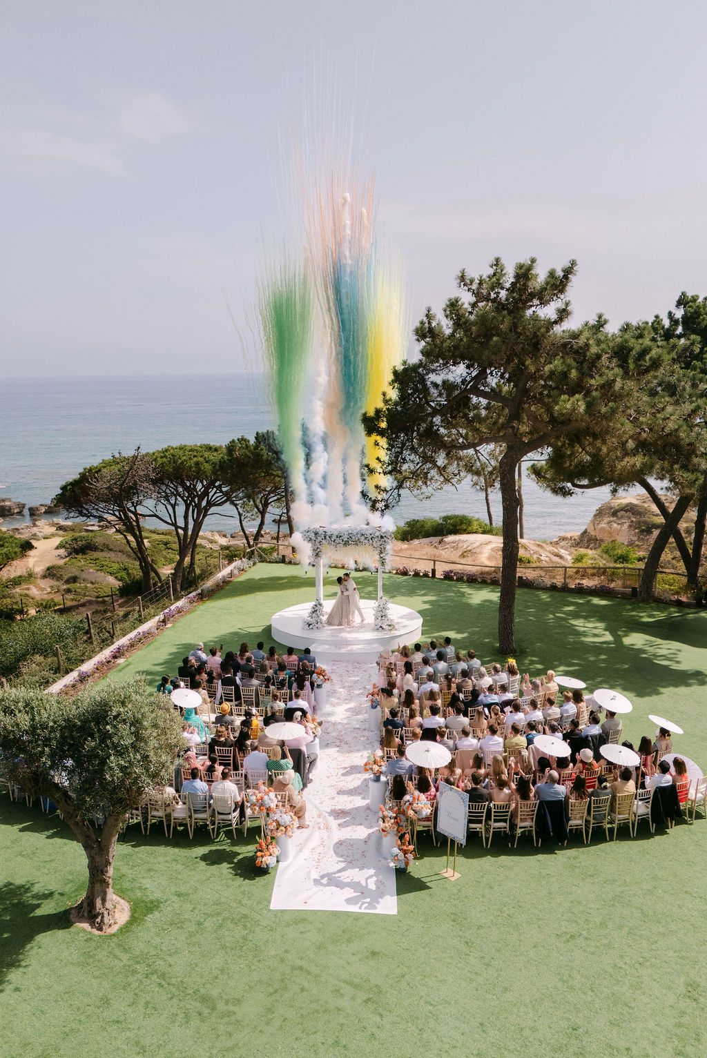 Outdoor wedding ceremony setup on a grassy oceanfront cliff with guests seated in a circular arrangement.