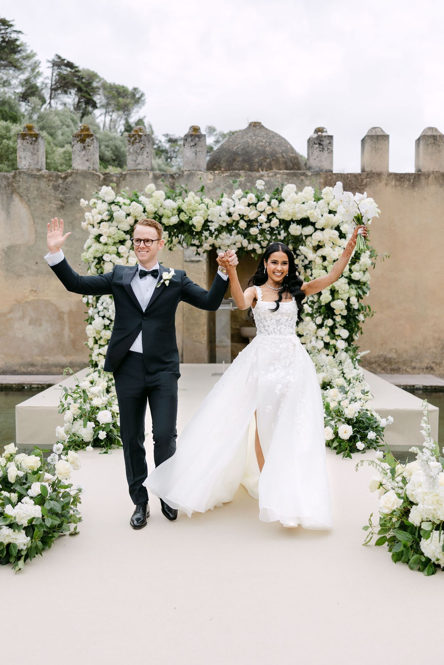 A joyful couple celebrating under an elegant white floral arch at their wedding ceremony.