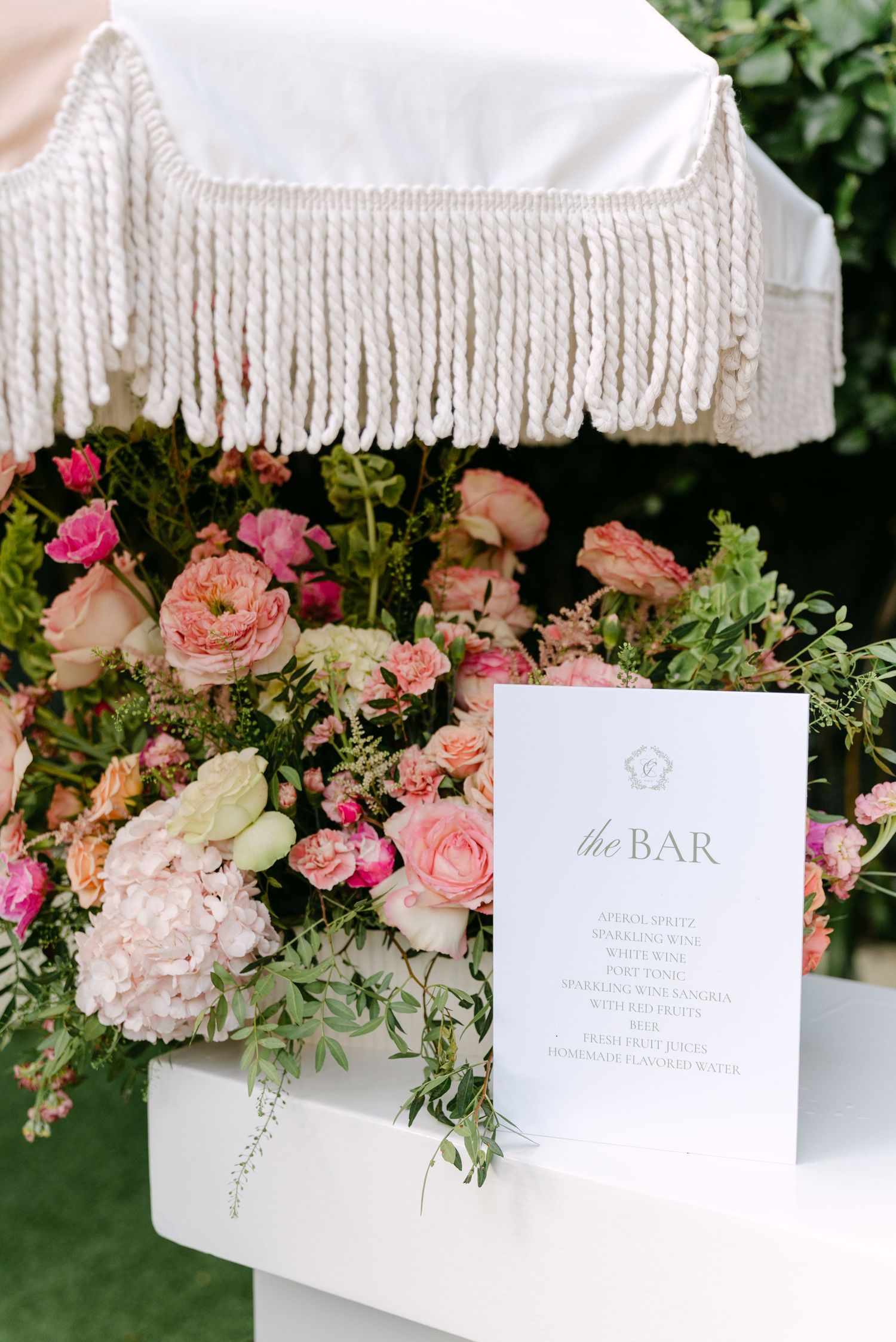 Elegant wedding bar setup with pink floral arrangements and white fringe decor against a green backdrop.