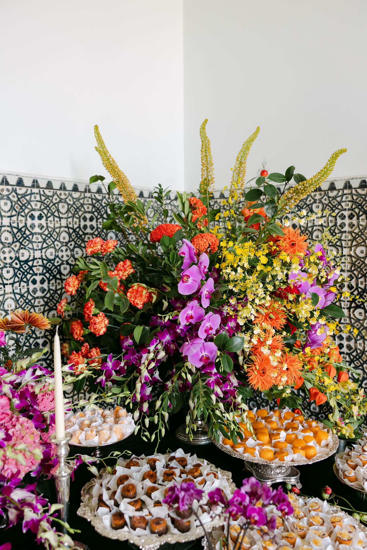 Vibrant floral arrangement with purple orchids and orange blooms alongside dessert display.