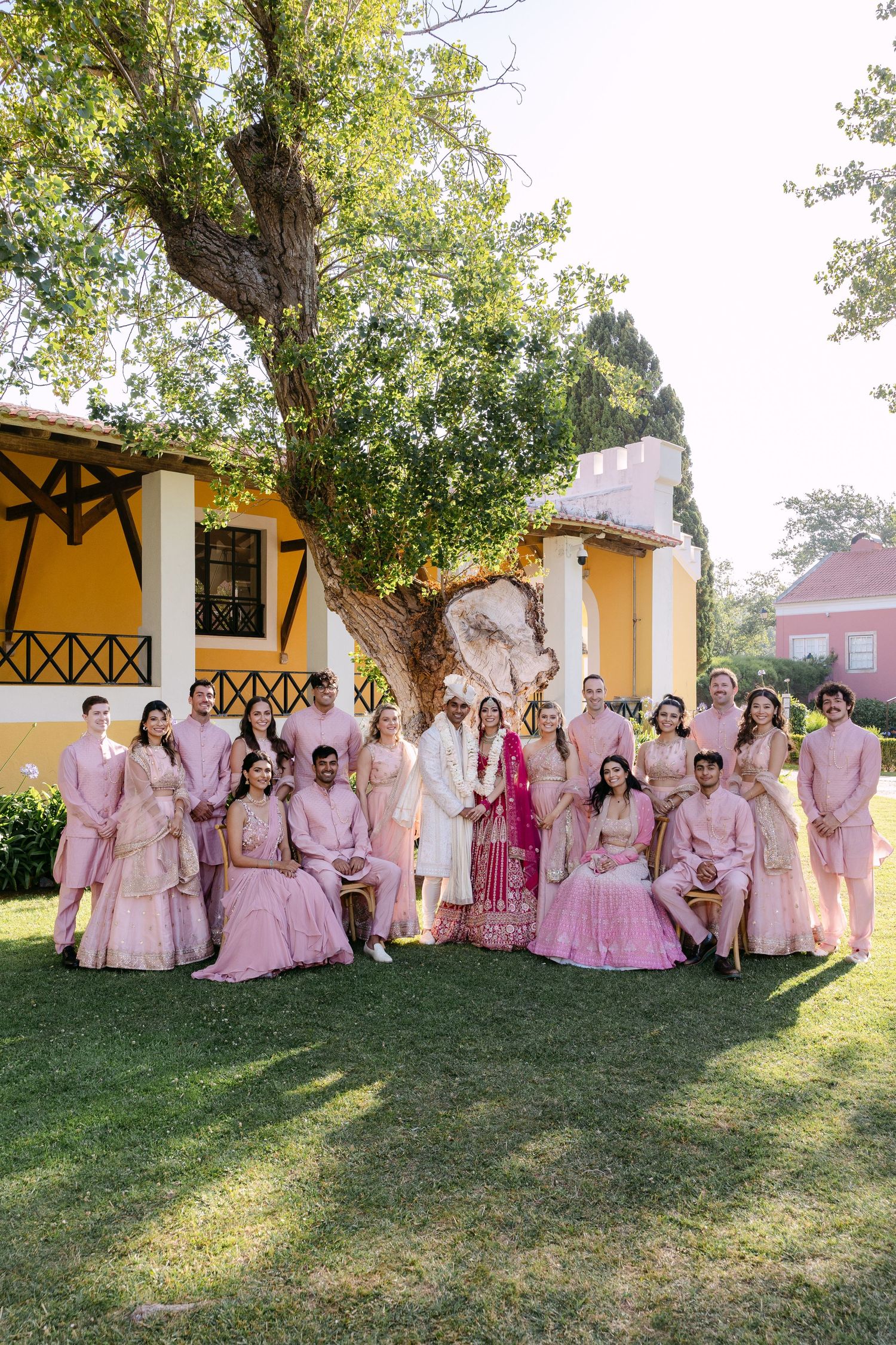 Large wedding party in pink and rose-colored outfits posing together on grassy lawn.