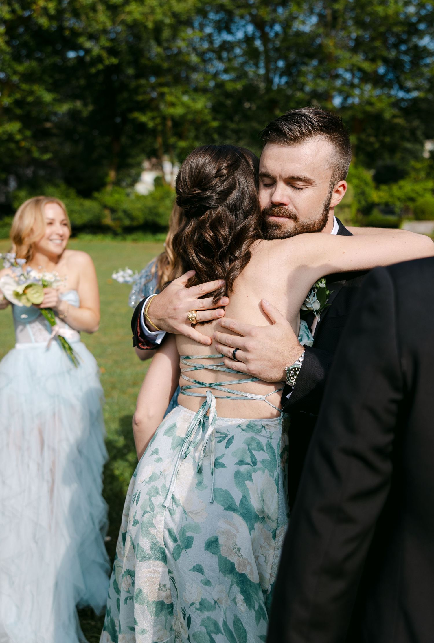 Wedding party in light blue dresses and black suits embracing outdoors in a garden setting.