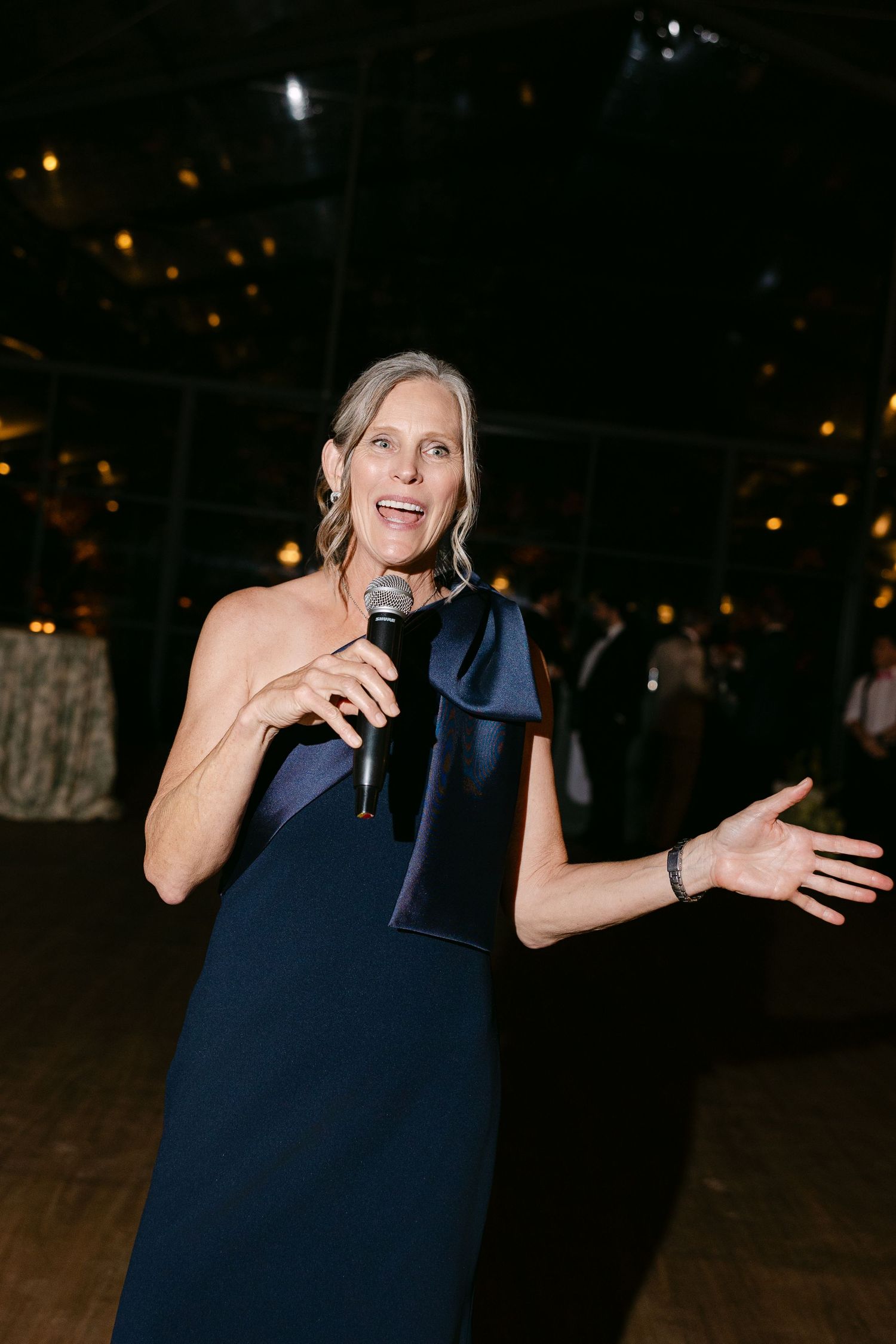 A person in a navy blue dress gestures expressively while dancing at an evening event.