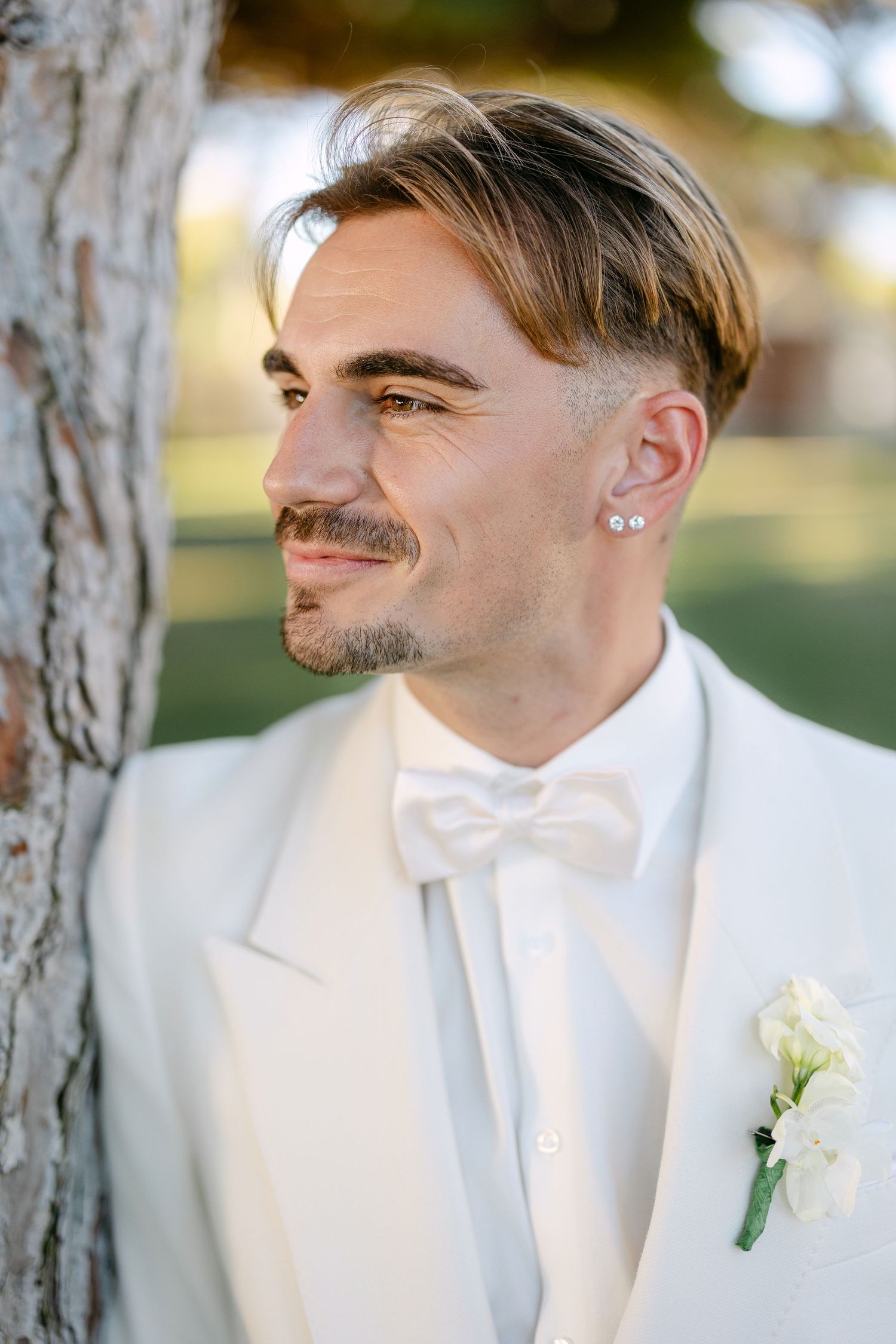 A groom in a white tuxedo with a boutonniere poses for portraits between trees.