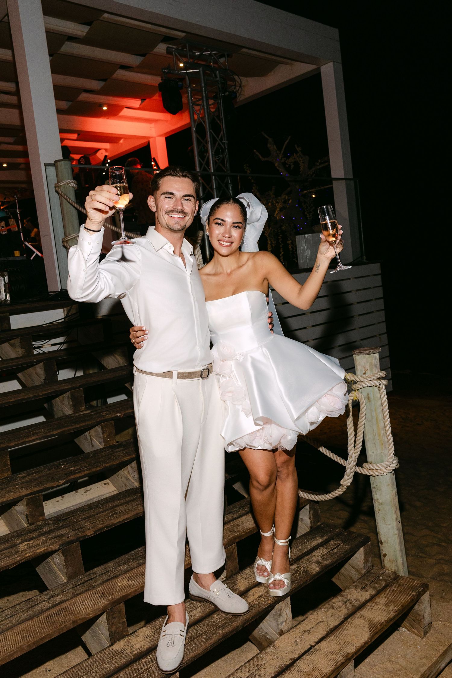 Newlyweds celebrating on wooden dock at night with festive lighting.