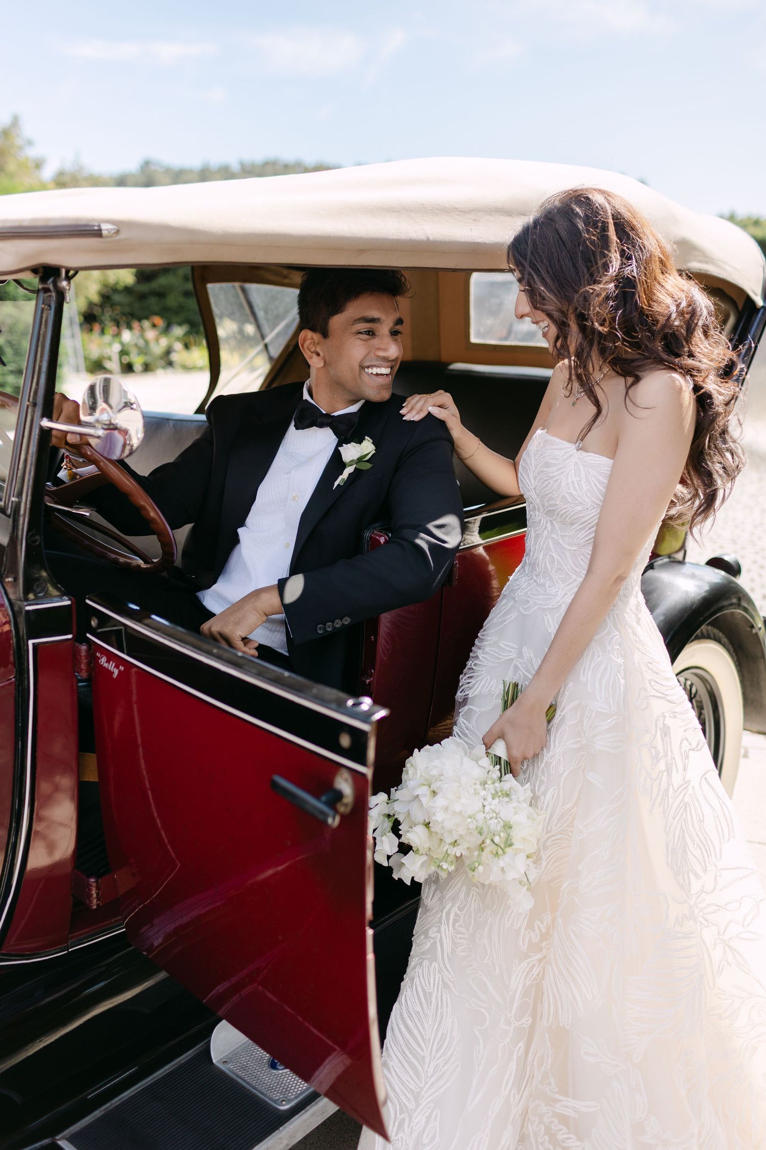 Wedding couple interacting near a classic vintage red car with elegant formal attire.