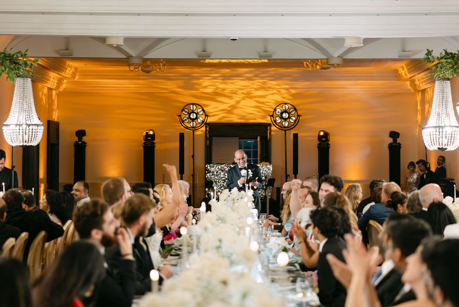 Wedding reception hall filled with guests and decorated with crystal chandeliers and floral arrangements.