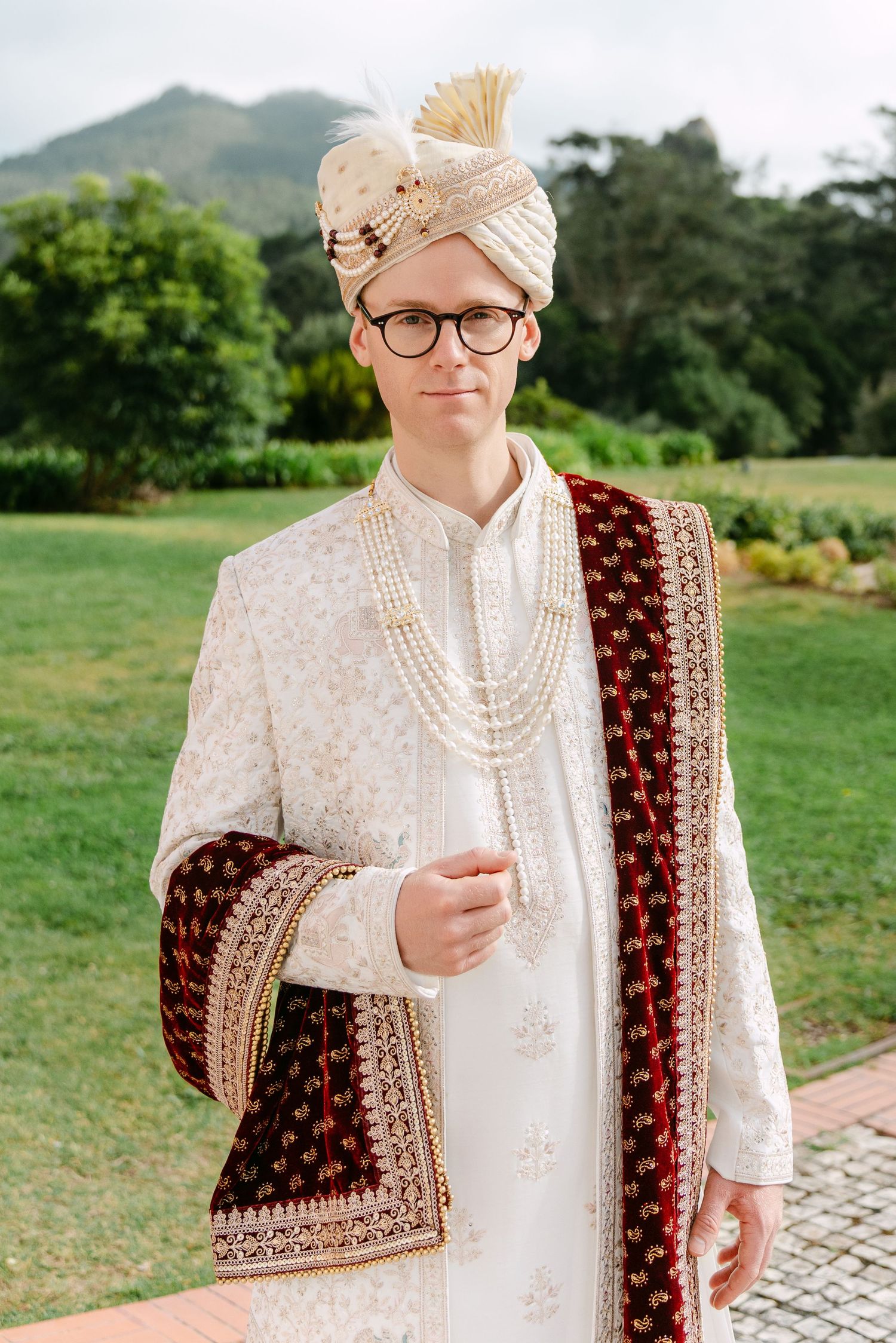 Groom in traditional cream sherwani with maroon velvet dupatta outdoors.