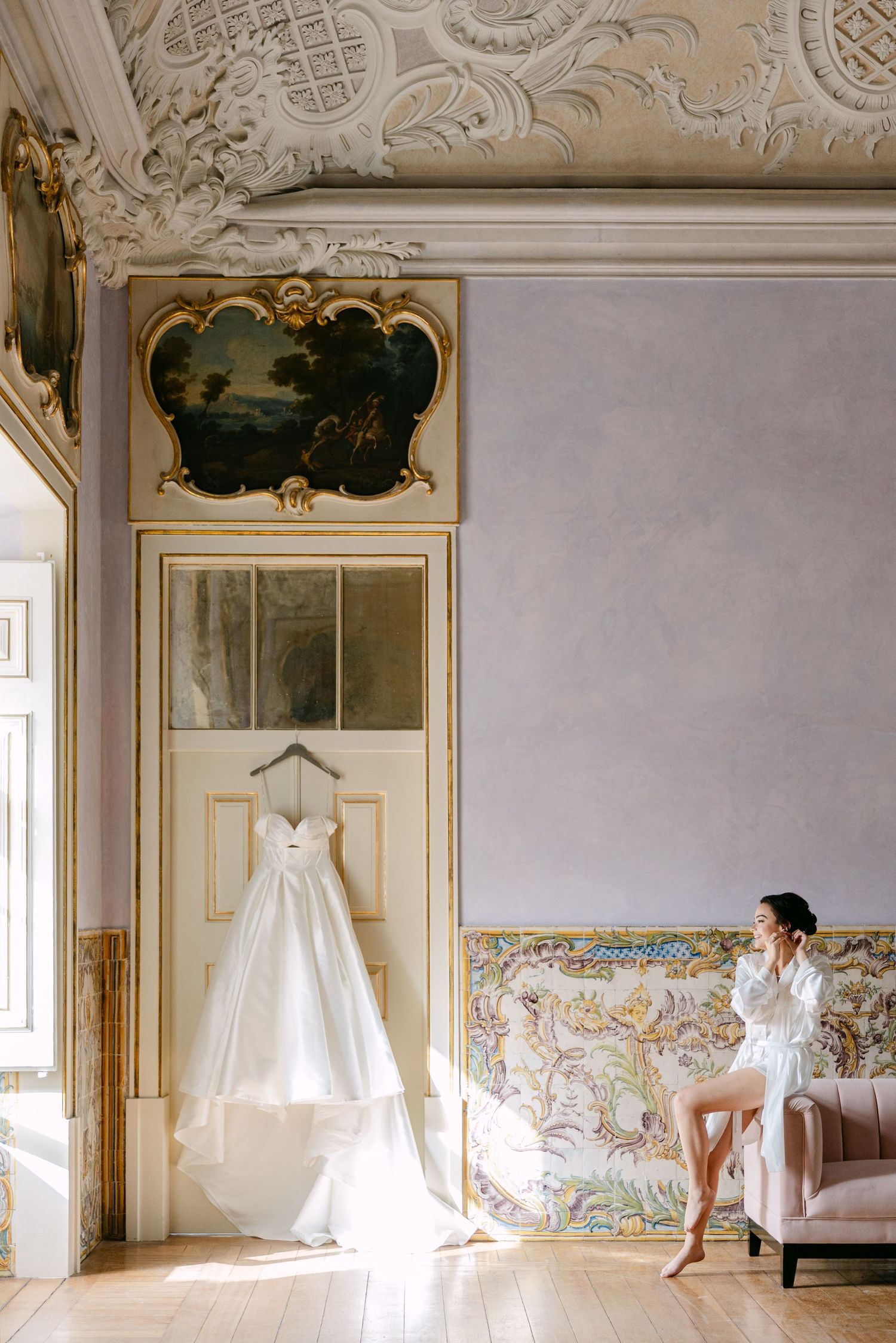 Elegant bridal gown hanging in ornate room with classical architectural details and natural light.