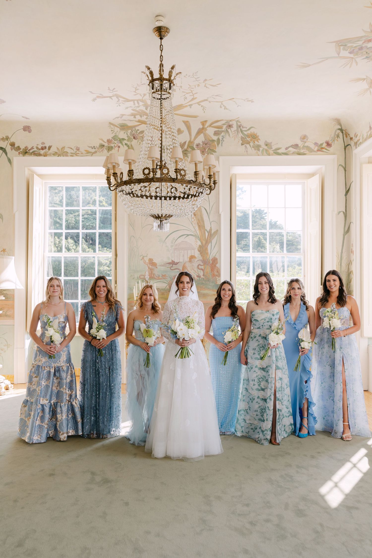 Wedding party in flowing blue and floral gowns posing in an elegant room with chandelier and ornate details.