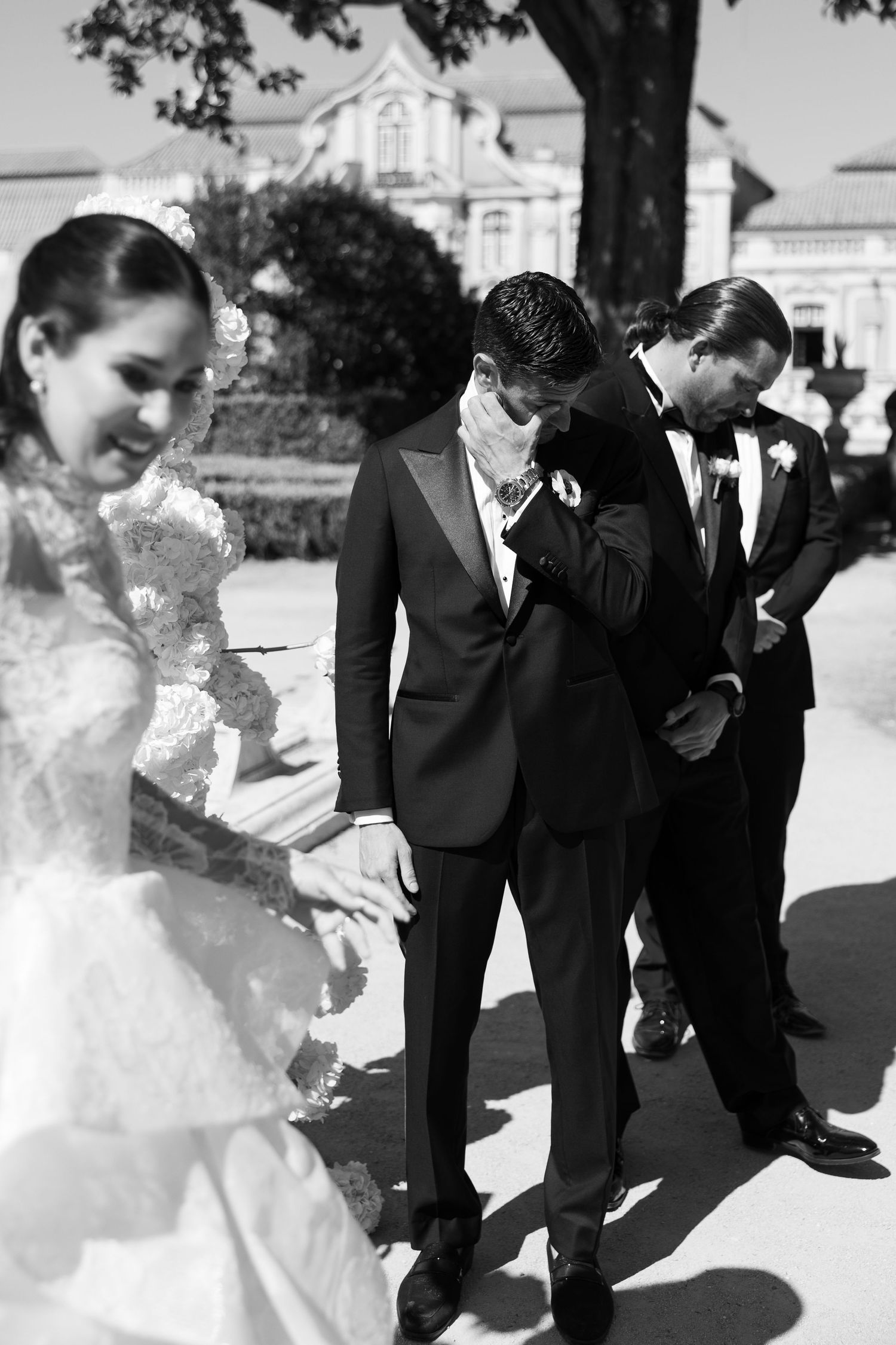 Wedding party in black suits and white dress laughing together outdoors on sunny day.
